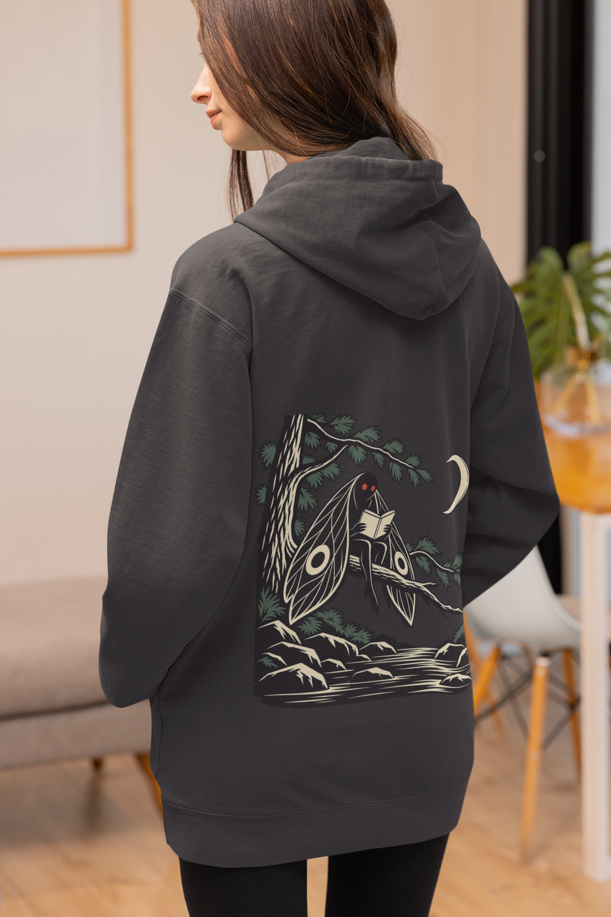 Night Library Midweight Hoodie in Black, worn by a woman seen from behind at home, showing the large print on the back. Digital product mockup.