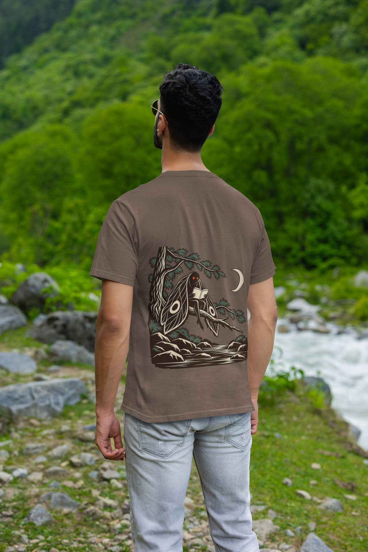 Night Library Garment-Dyed Tee in Espresso, worn by a man seen from behind by a mountain river, showing the large print on the back. Digital product mockup.