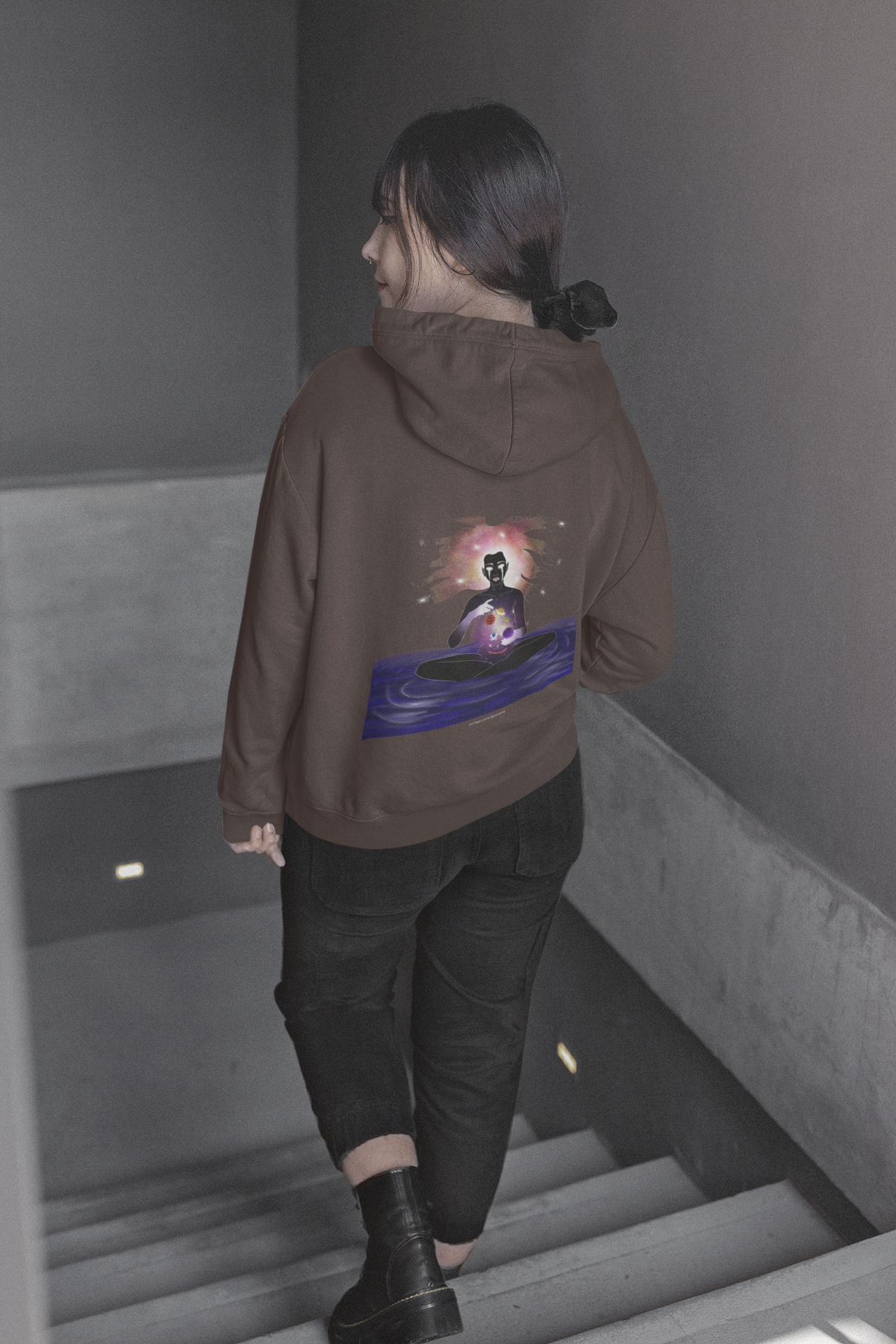 Orbit Keeper Heavyweight Hoodie in Brown, worn by a woman seen from behind walking down a staircase, showing the large print on the back. Digital product mockup.