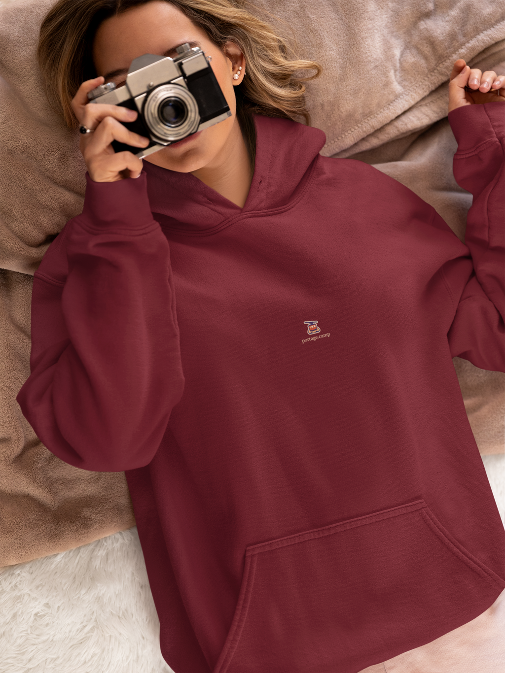 Cloud Ferry Midweight Hoodie in Maroon, worn by a woman in bed with a film camera, with the Cloud Ferry logo and portage.camp on the chest. Digital product mockup.