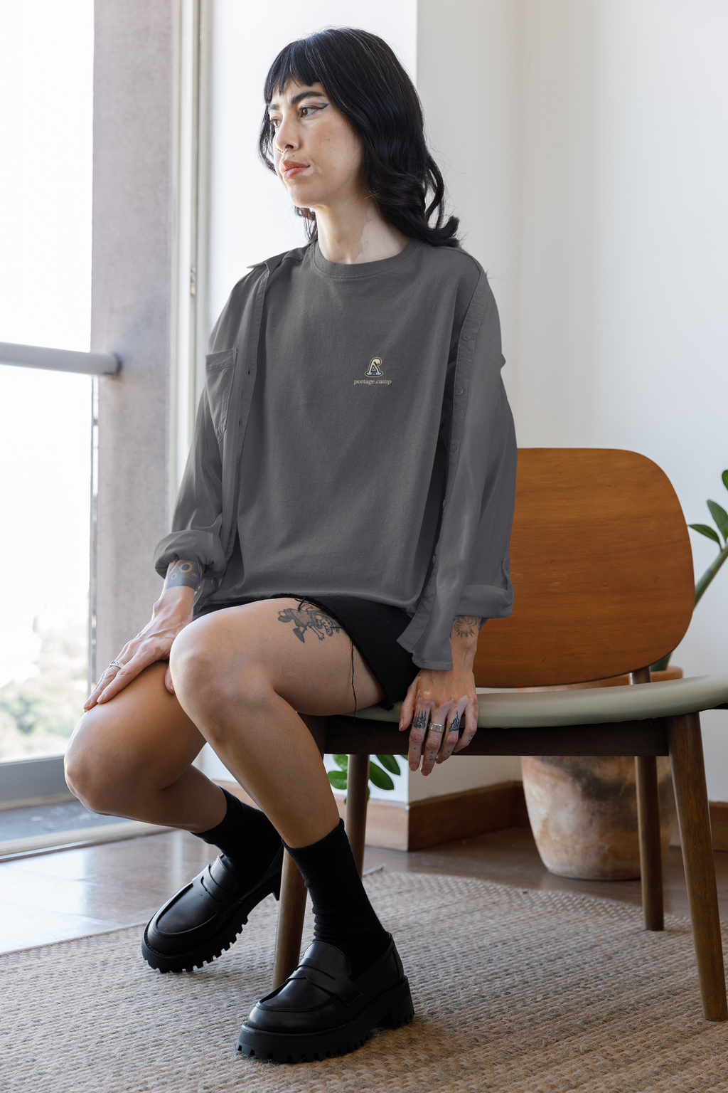 Small Hours Garment-Dyed Tee in Graphite, worn by a woman seated by a window at home, with the Small Hours logo and portage.camp on the chest. Digital product mockup.