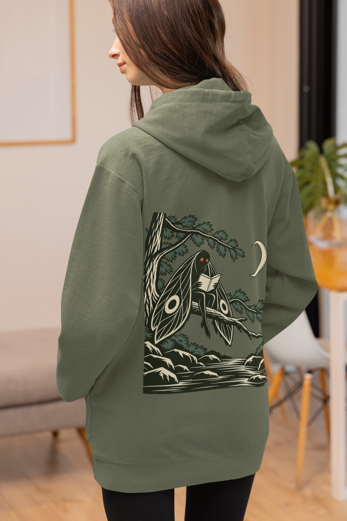 Night Library Zip Hoodie in Military Green, worn by a woman seen from behind at home, showing the large print on the back. Digital product mockup.