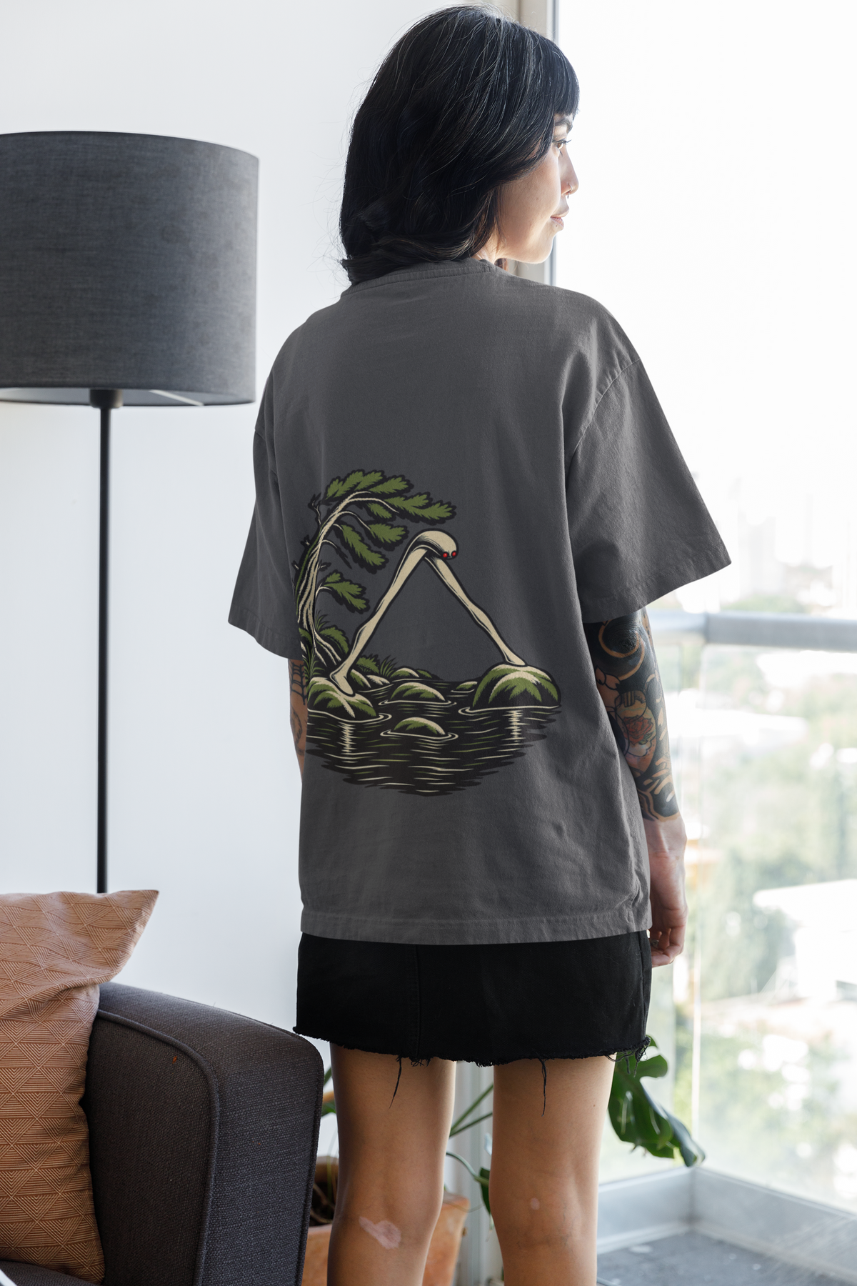 Small Hours Garment-Dyed Tee in Graphite, worn by a woman seen from behind at home by a bright window, showing the large print on the back. Digital product mockup.