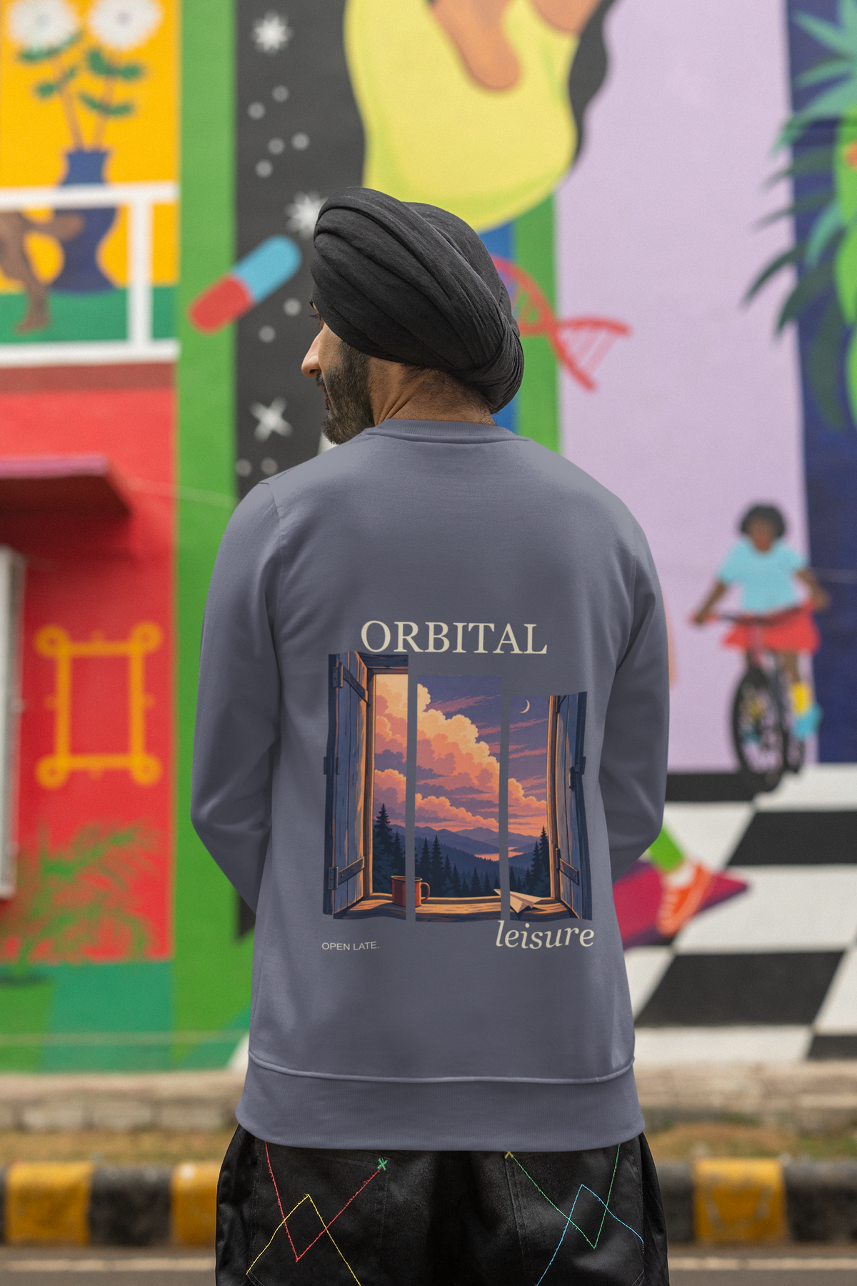 Open Late Garment-Dyed Crewneck in Denim, worn by a man seen from behind in a turban by a painted mural, showing the large print on the back. Digital product mockup.