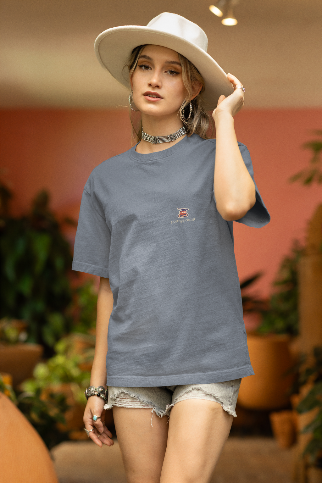Cloud Ferry Garment-Dyed Tee in Blue Jean, worn by a woman in a sun hat among potted plants, with the Cloud Ferry logo and portage.camp on the chest. Digital product mockup.