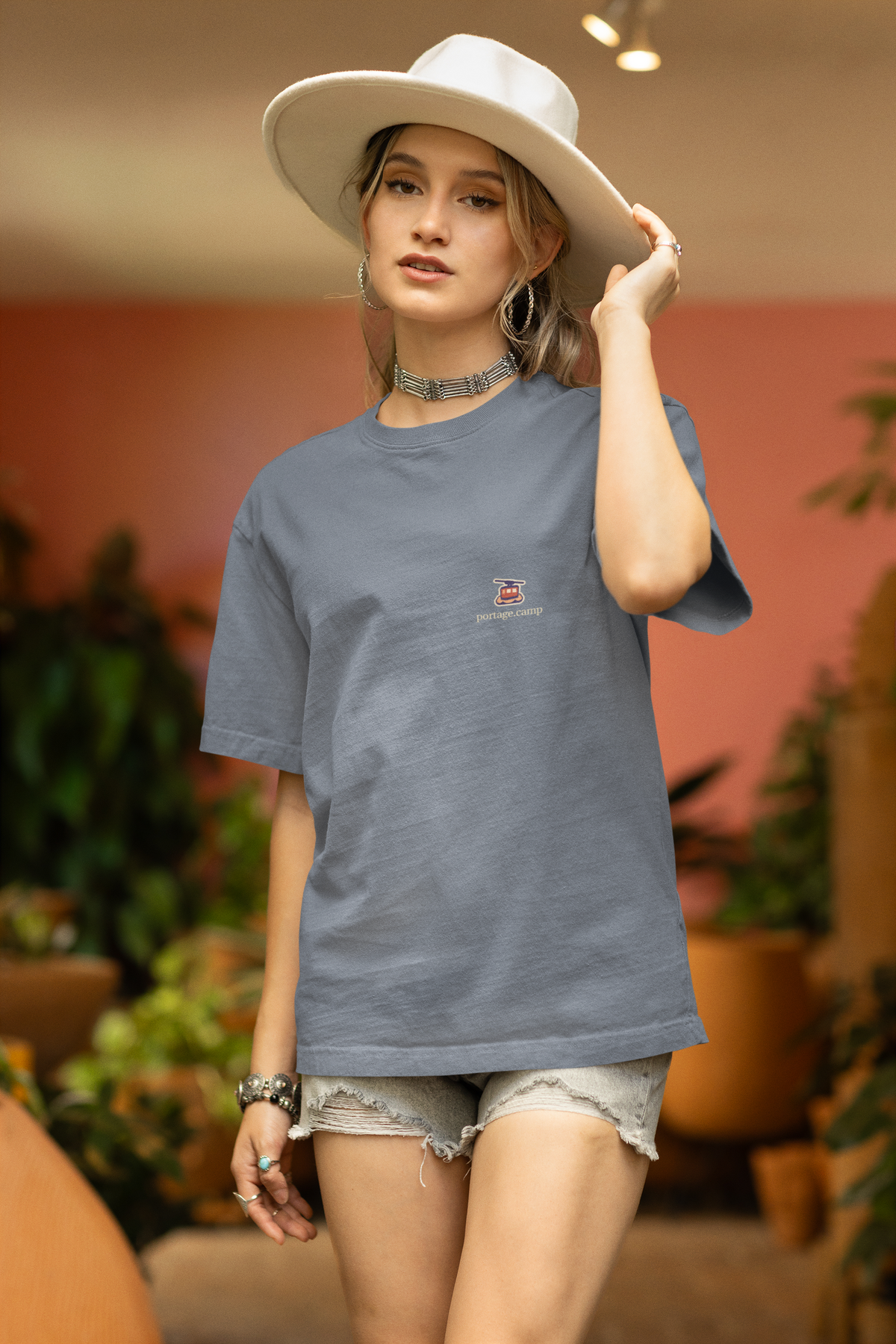 Cloud Ferry Garment-Dyed Tee in Blue Jean, worn by a woman in a sun hat among potted plants, with the Cloud Ferry logo and portage.camp on the chest. Digital product mockup.