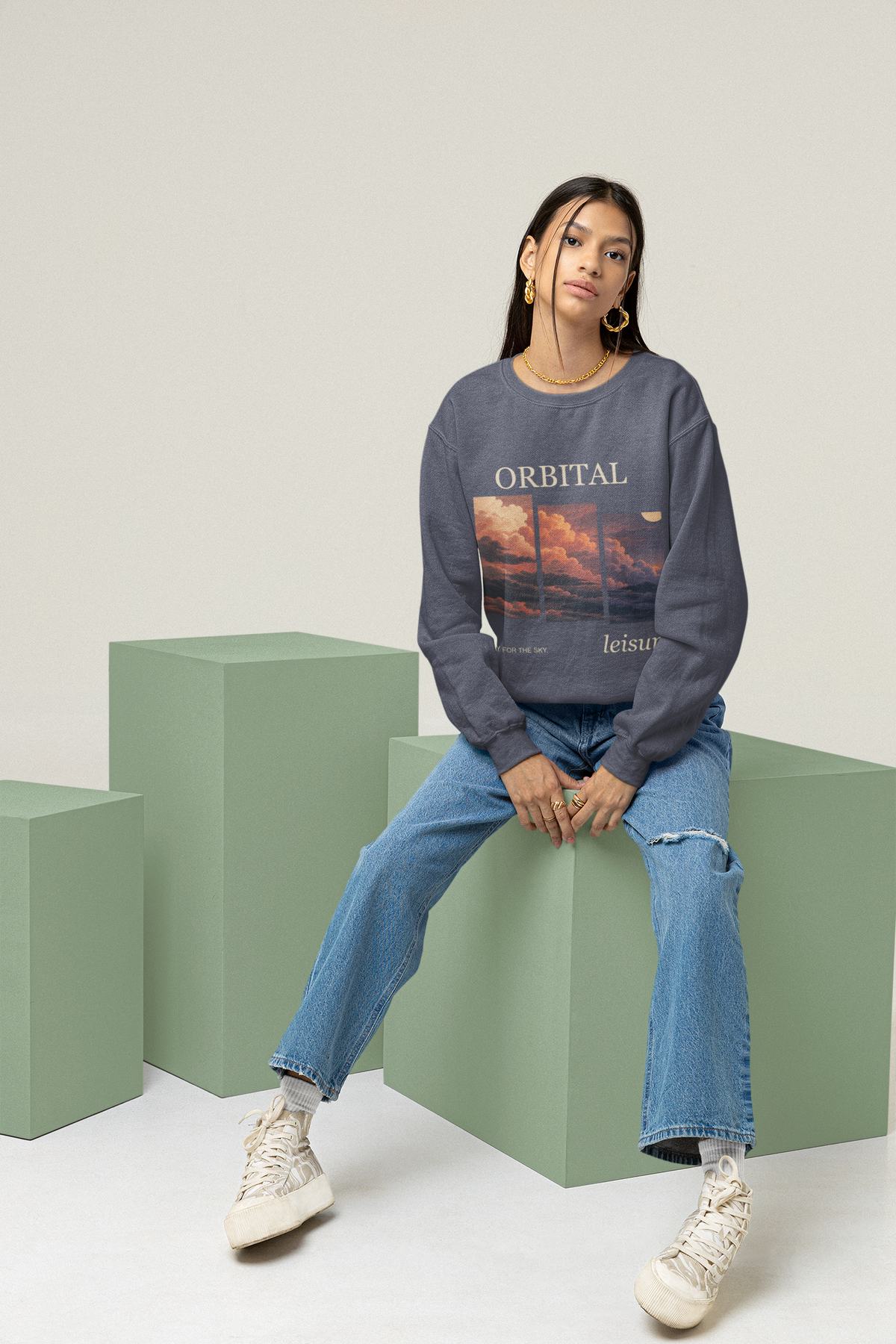 Stay for the Sky Garment-Dyed Crewneck (Front Print) in Denim, worn by a woman seated on green studio blocks, showing the large print on the front. Digital product mockup.