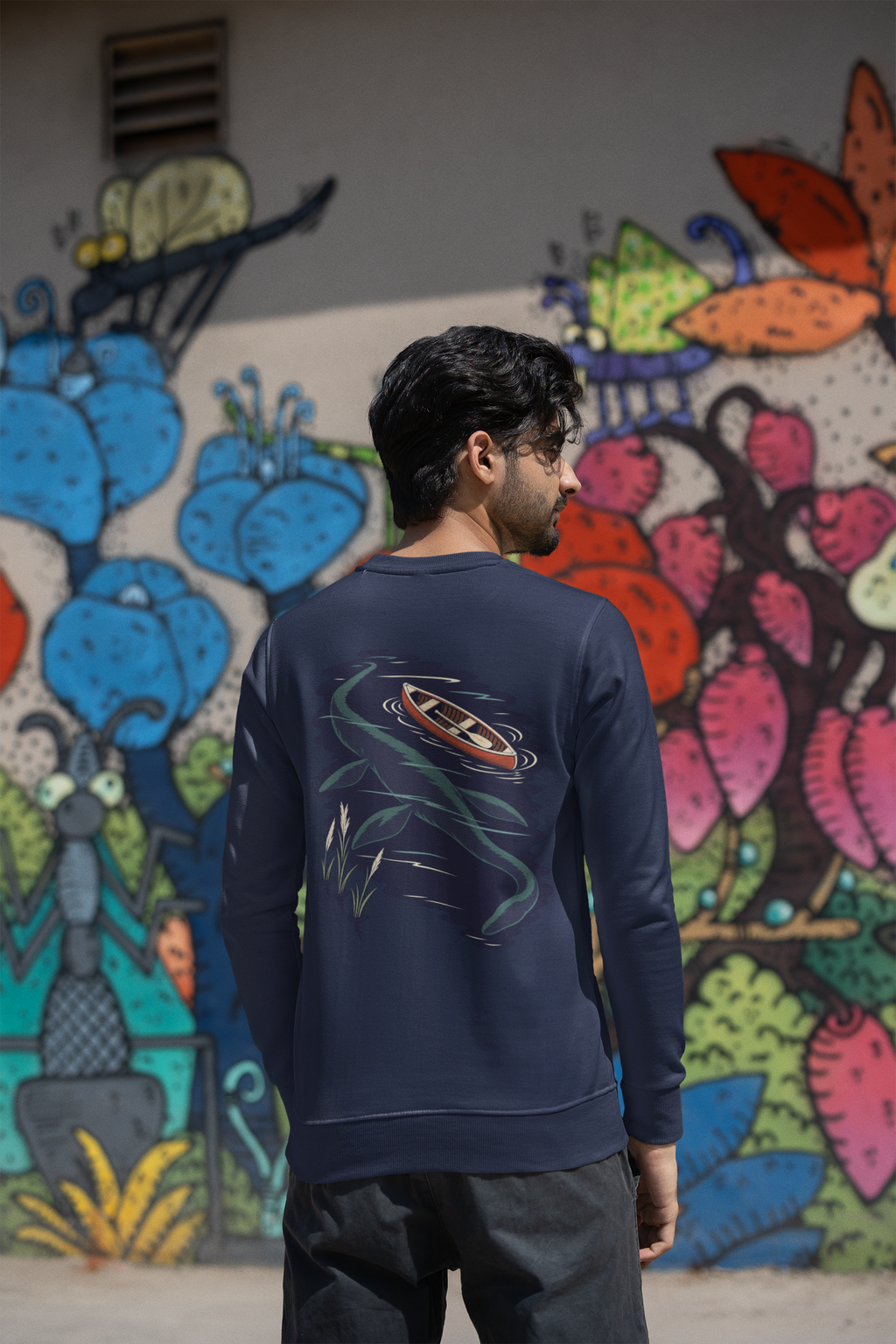 Seen Briefly Garment-Dyed Crewneck in True Navy, worn by a man seen from behind by a painted wall in the city, showing the large print on the back. Digital product mockup.