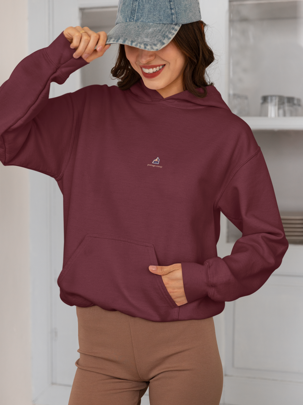 A Different Sky Midweight Hoodie in Maroon, worn by a woman smiling in a kitchen, with the A Different Sky logo and portage.camp on the chest. Digital product mockup.