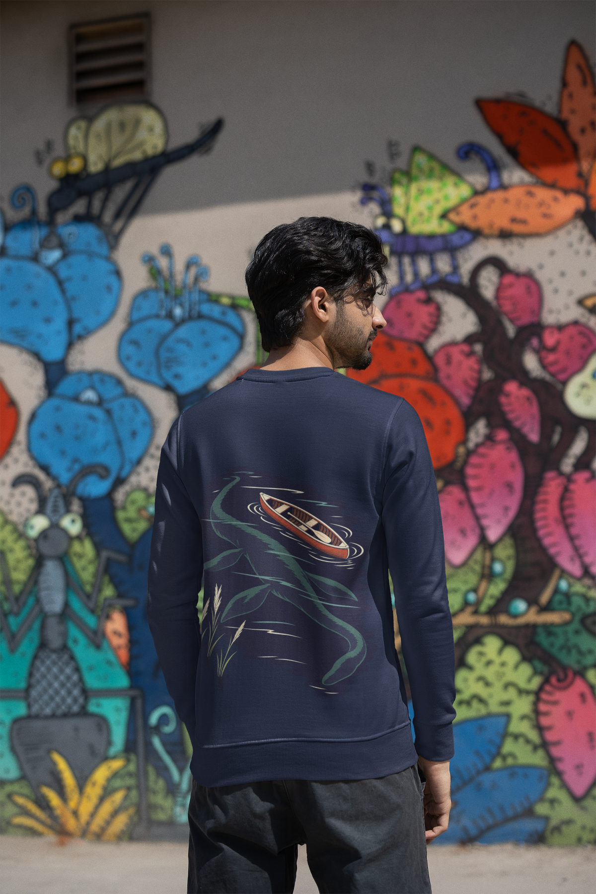 Seen Briefly Garment-Dyed Crewneck in True Navy, worn by a man seen from behind by a painted wall in the city, showing the large print on the back. Digital product mockup.