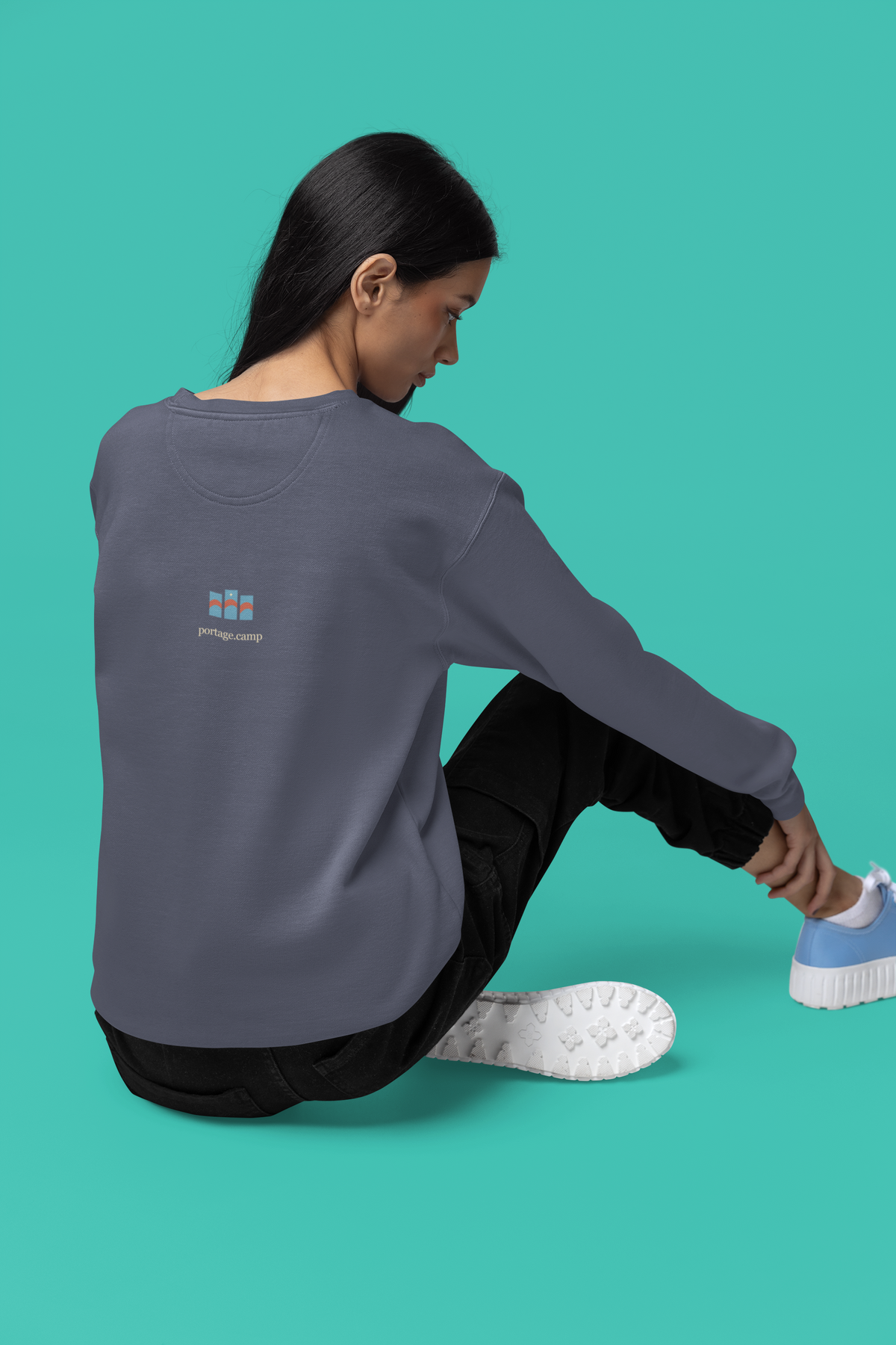 Stay for the Sky Garment-Dyed Crewneck (Front Print) in Denim, worn by a woman seen from behind seated on a studio floor, showing the portage.camp emblem at the upper back. Digital product mockup.