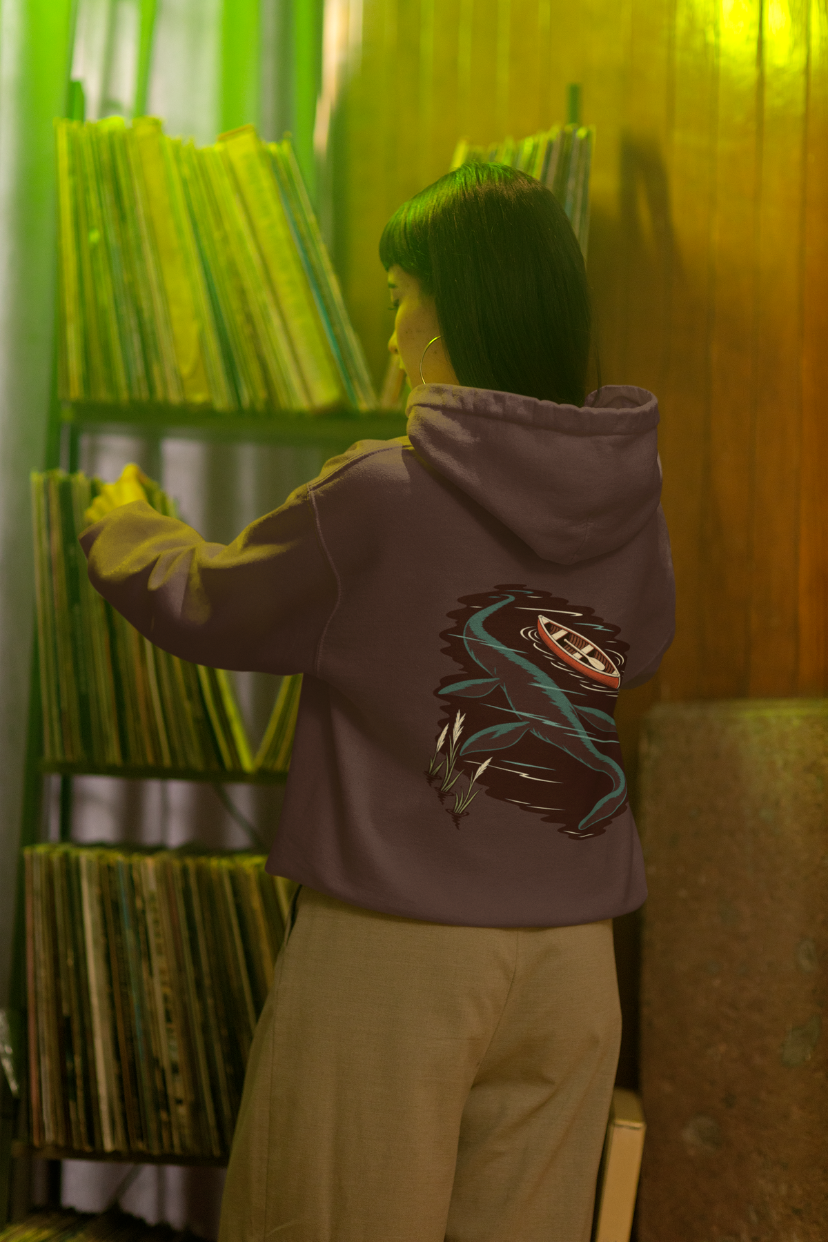 Seen Briefly Heavyweight Hoodie in Brown, worn by a woman seen from behind in a vintage record shop, showing the large print on the back. Digital product mockup.