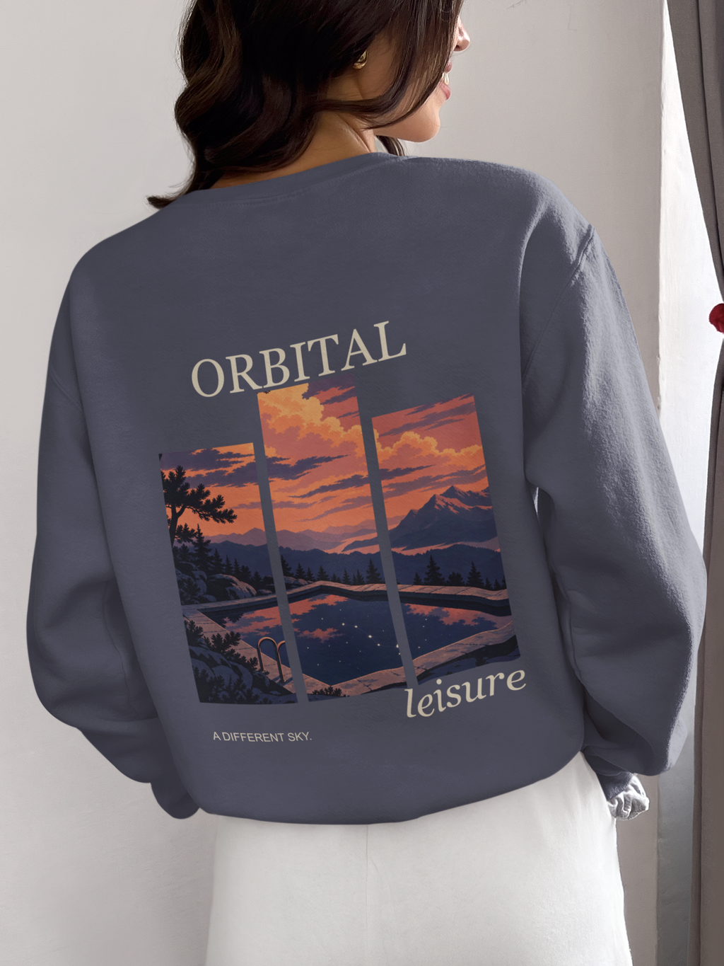 A Different Sky Garment-Dyed Crewneck in Denim, worn by a woman seen from behind at home, showing the large print on the back. Digital product mockup.
