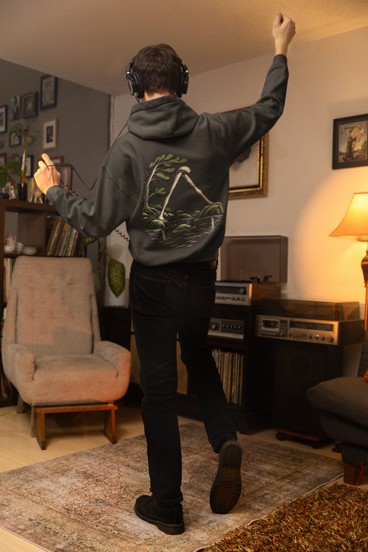 Small Hours Zip Hoodie in Black, worn by a man seen from behind in a living room at night, showing the large print on the back. Digital product mockup.
