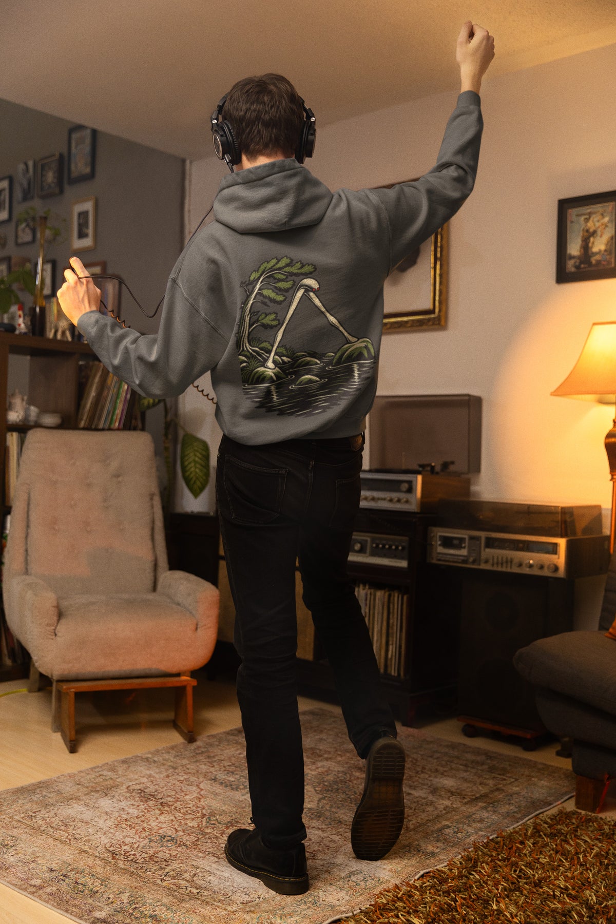Small Hours Zip Hoodie in Dark Heather, worn by a man seen from behind in a living room at night, showing the large print on the back. Digital product mockup.