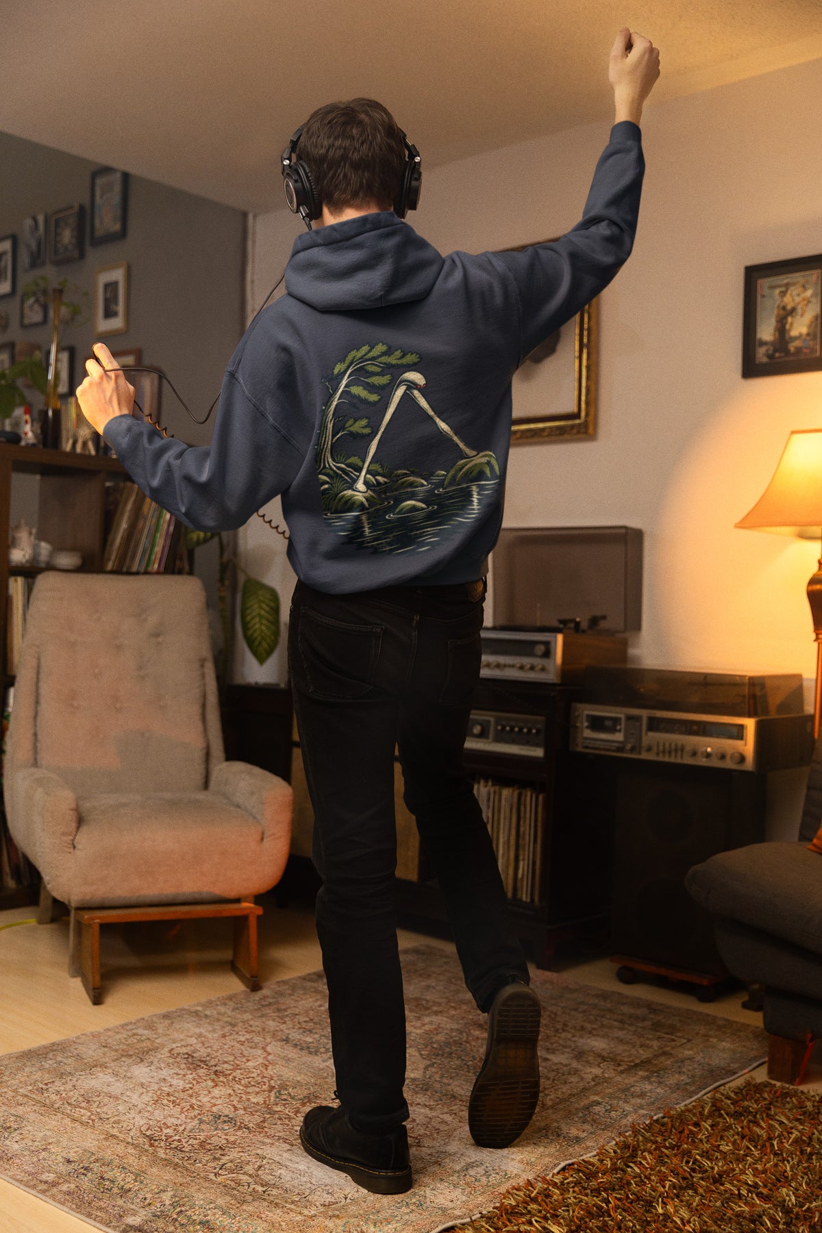Small Hours Zip Hoodie in Navy, worn by a man seen from behind in a living room at night, showing the large print on the back. Digital product mockup.