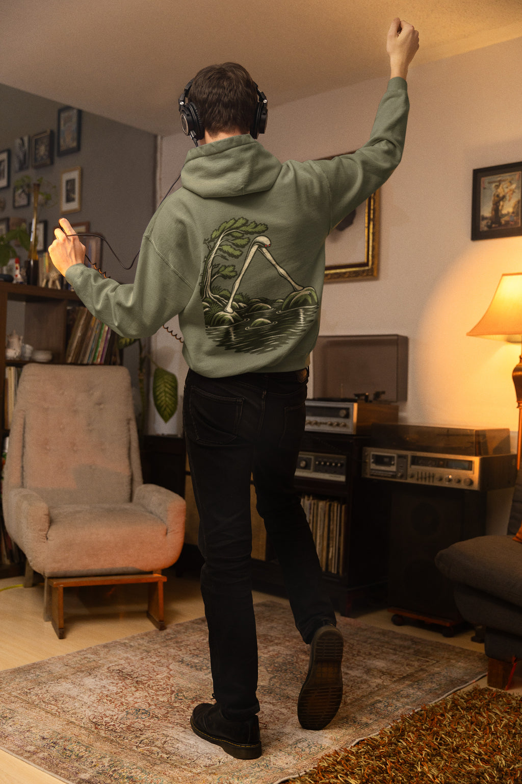 Small Hours Zip Hoodie in Military Green, worn by a man seen from behind in a living room at night, showing the large print on the back. Digital product mockup.