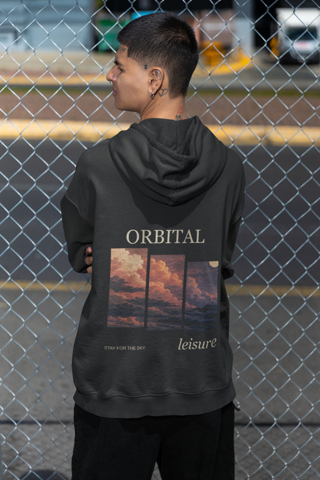 Stay for the Sky Zip Hoodie in Black, worn by a man seen from behind by a chain-link fence, showing the large print on the back. Digital product mockup.