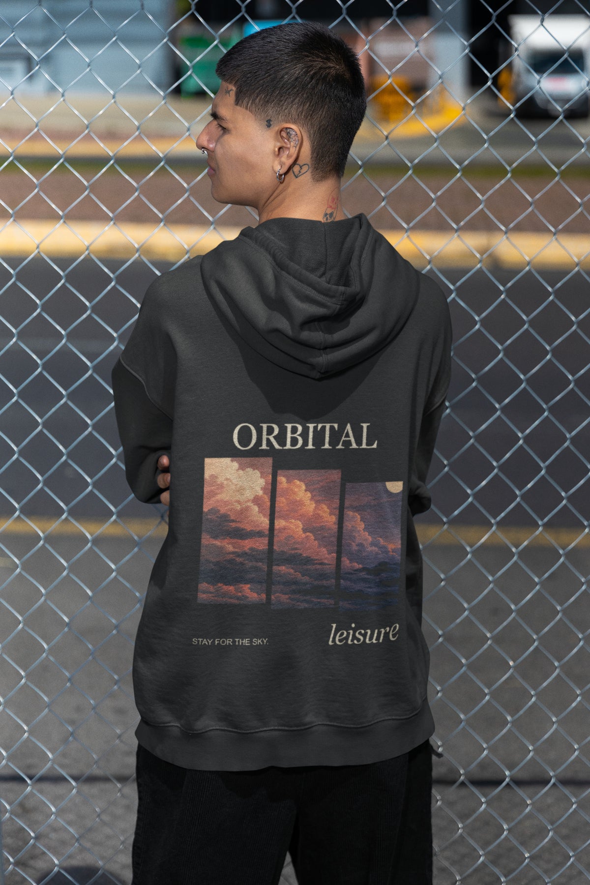 Stay for the Sky Zip Hoodie in Black, worn by a man seen from behind by a chain-link fence, showing the large print on the back. Digital product mockup.