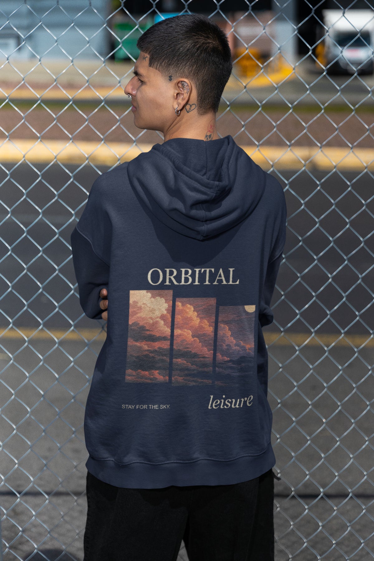 Stay for the Sky Zip Hoodie in Navy, worn by a man seen from behind by a chain-link fence, showing the large print on the back. Digital product mockup.