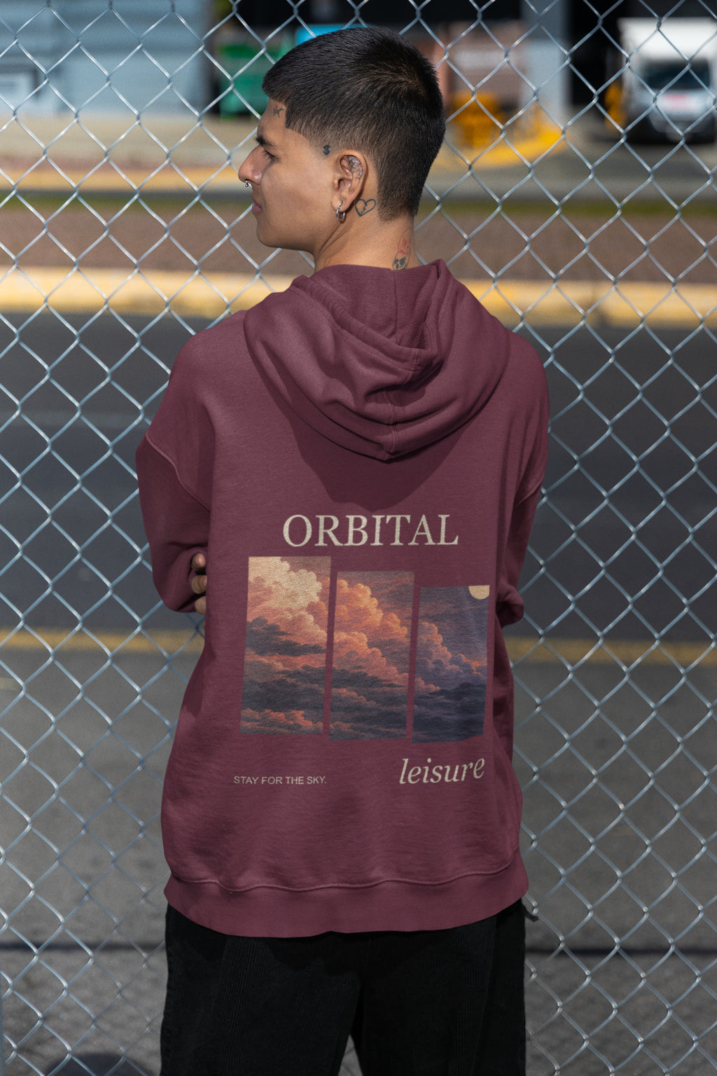 Stay for the Sky Zip Hoodie in Maroon, worn by a man seen from behind by a chain-link fence, showing the large print on the back. Digital product mockup.