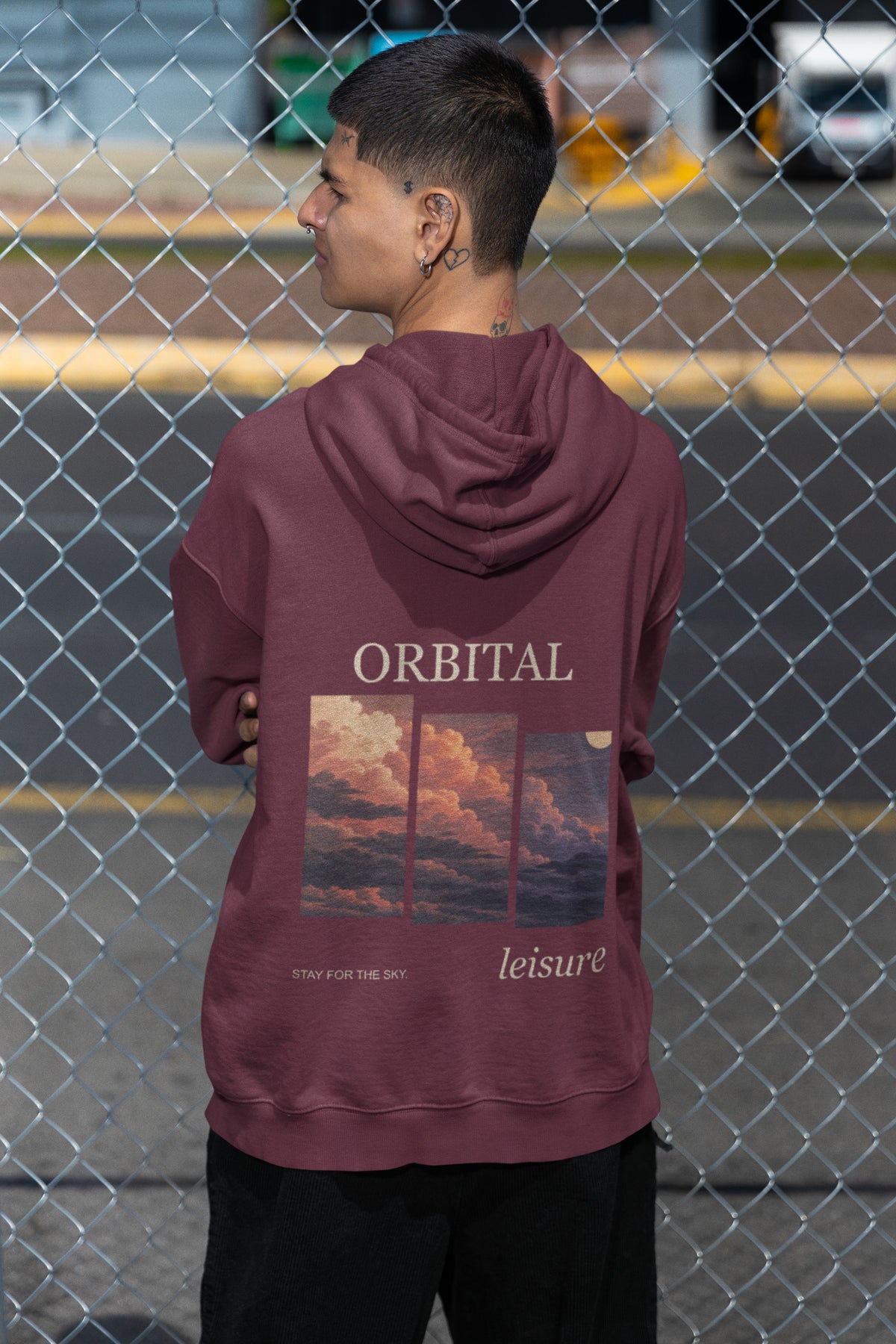 Stay for the Sky Zip Hoodie in Maroon, worn by a man seen from behind by a chain-link fence, showing the large print on the back. Digital product mockup.