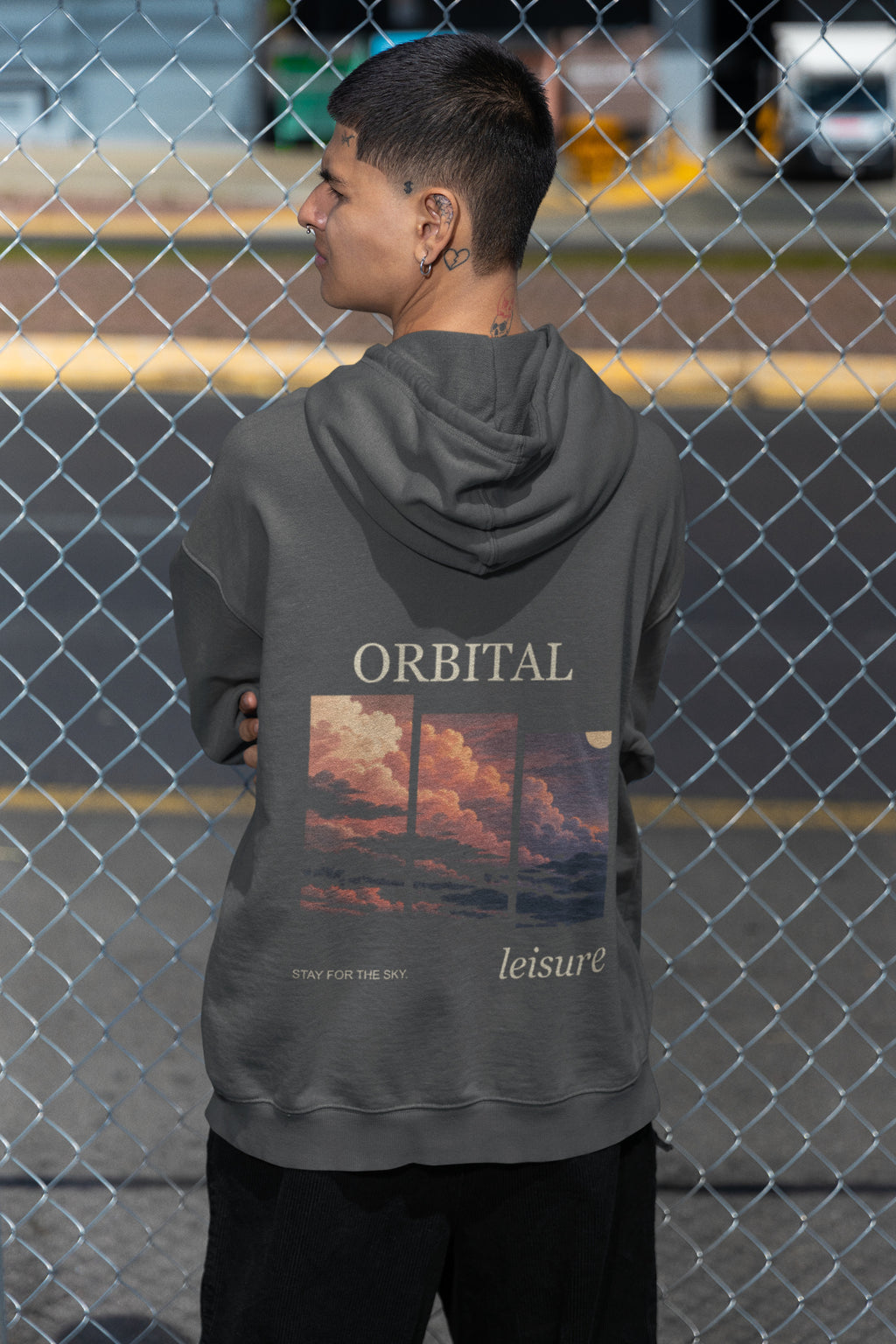Stay for the Sky Zip Hoodie in Dark Heather, worn by a man seen from behind by a chain-link fence, showing the large print on the back. Digital product mockup.