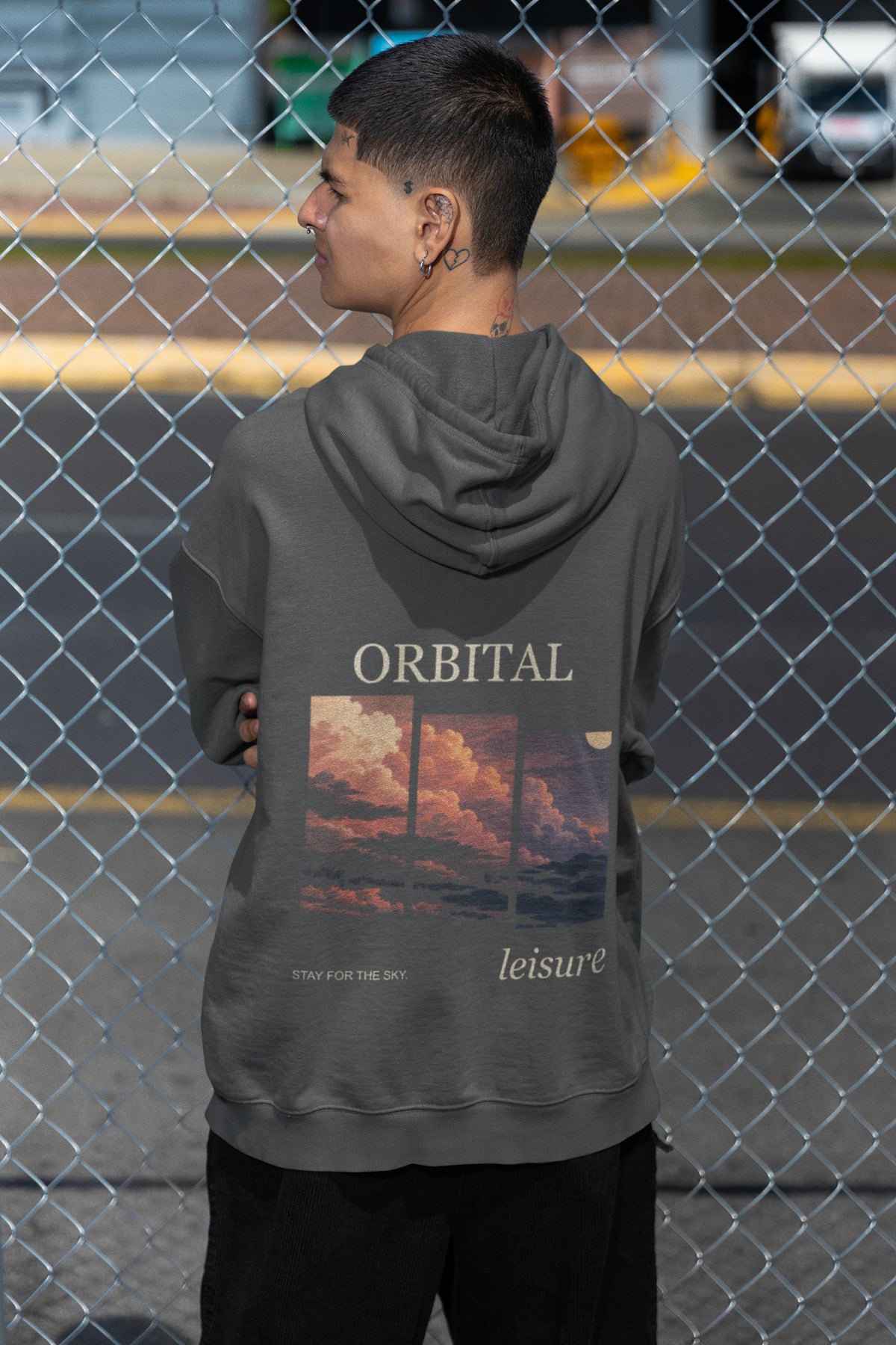 Stay for the Sky Zip Hoodie in Dark Heather, worn by a man seen from behind by a chain-link fence, showing the large print on the back. Digital product mockup.