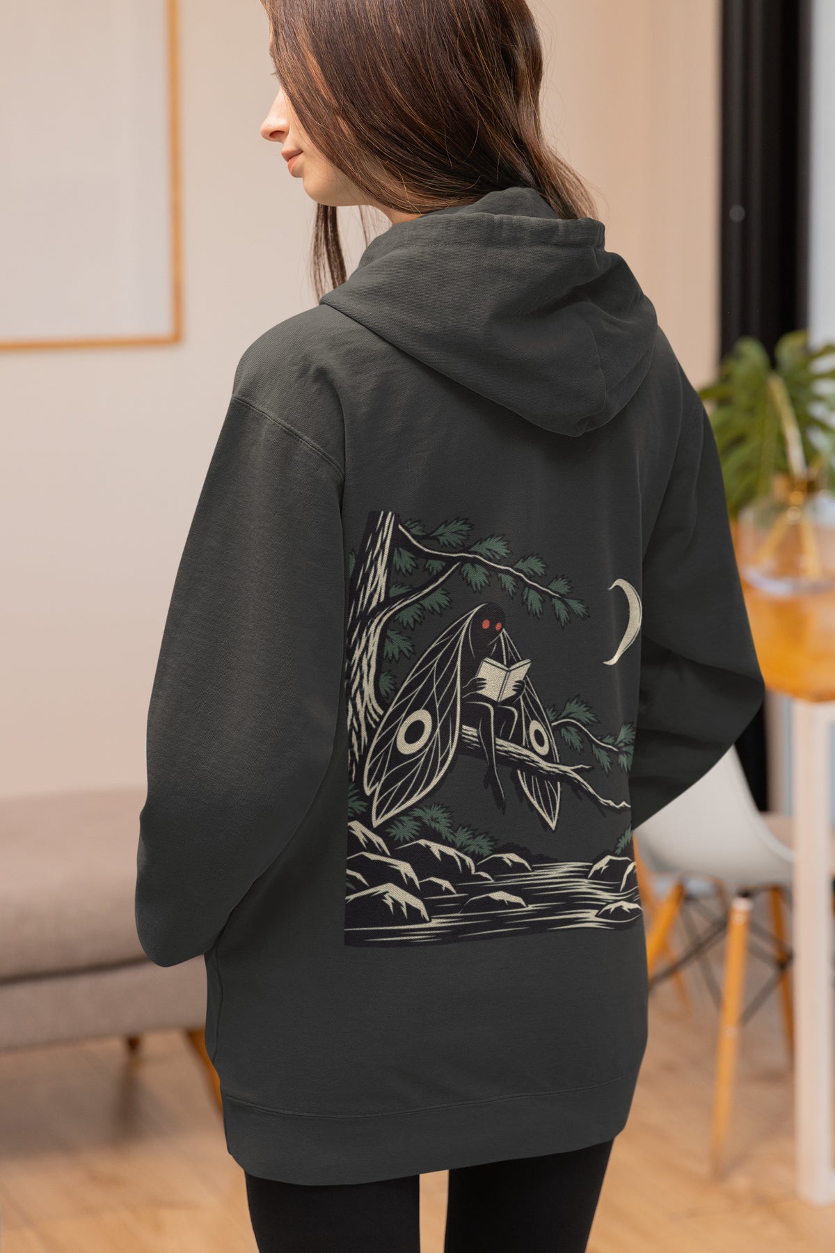 Night Library Zip Hoodie in Black, worn by a woman seen from behind at home, showing the large print on the back. Digital product mockup.