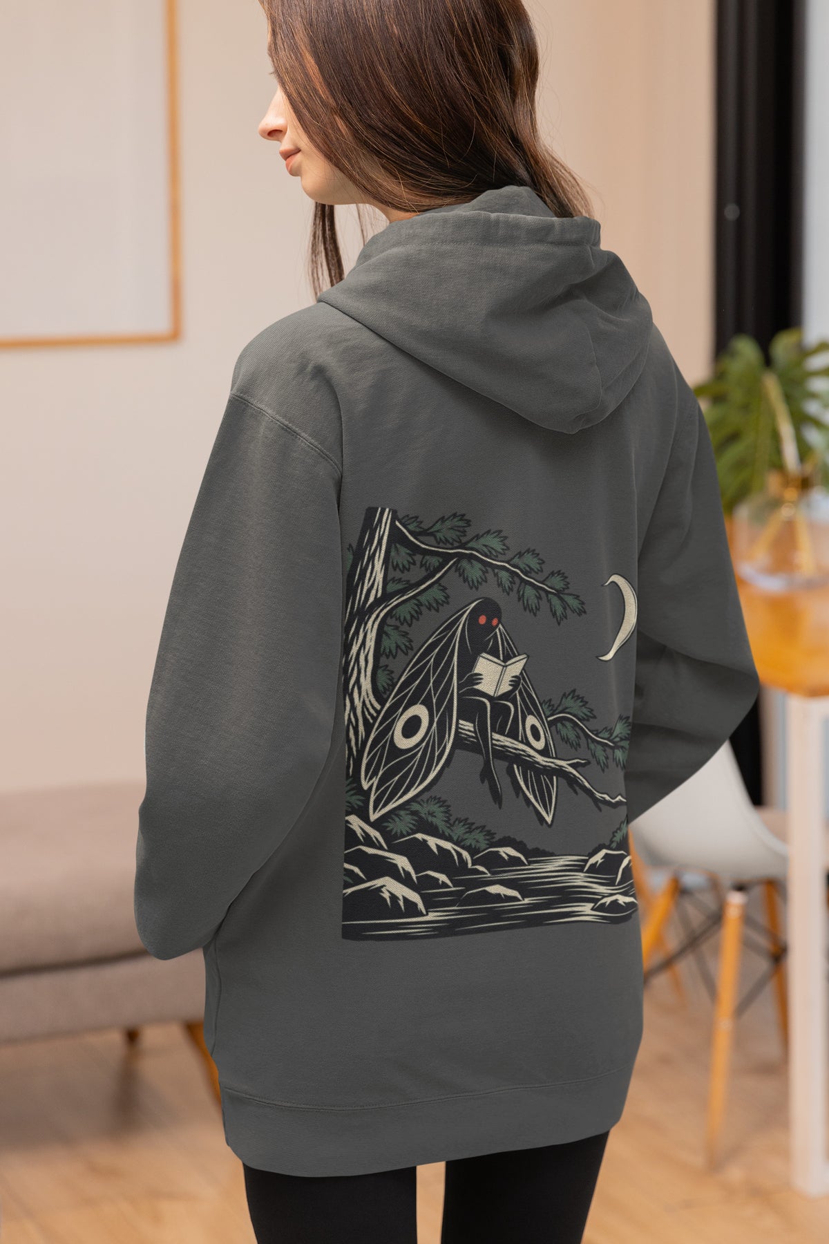Night Library Zip Hoodie in Dark Heather, worn by a woman seen from behind at home, showing the large print on the back. Digital product mockup.