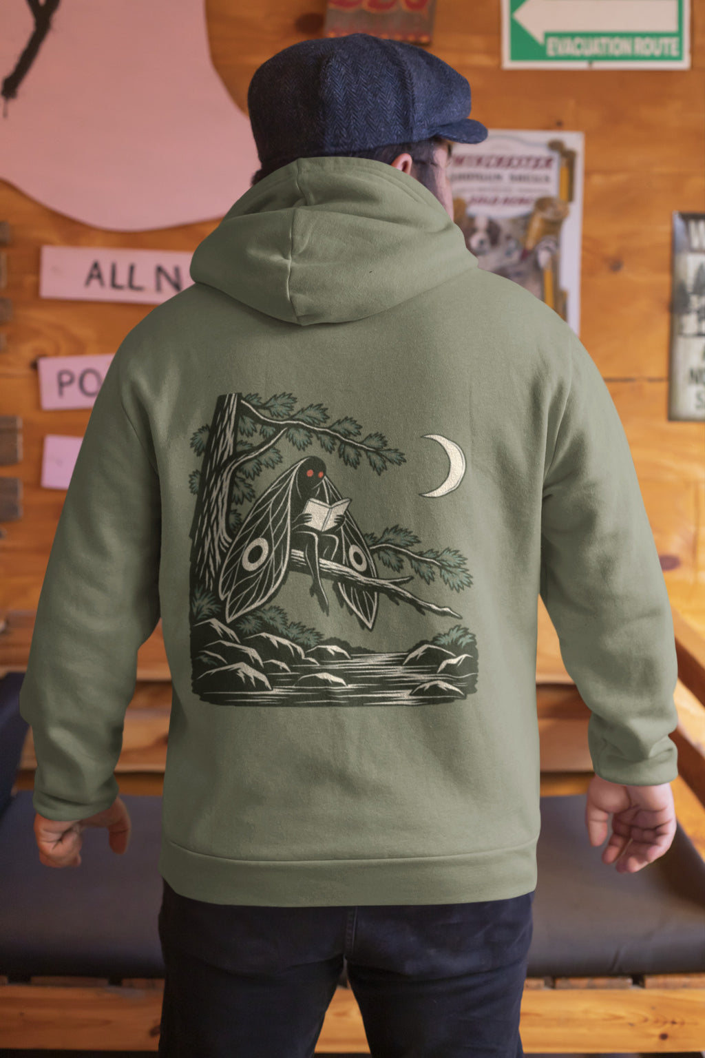 Night Library Zip Hoodie in Military Green, worn by a man seen from behind in a wood-panelled bar, showing the large print on the back. Digital product mockup.