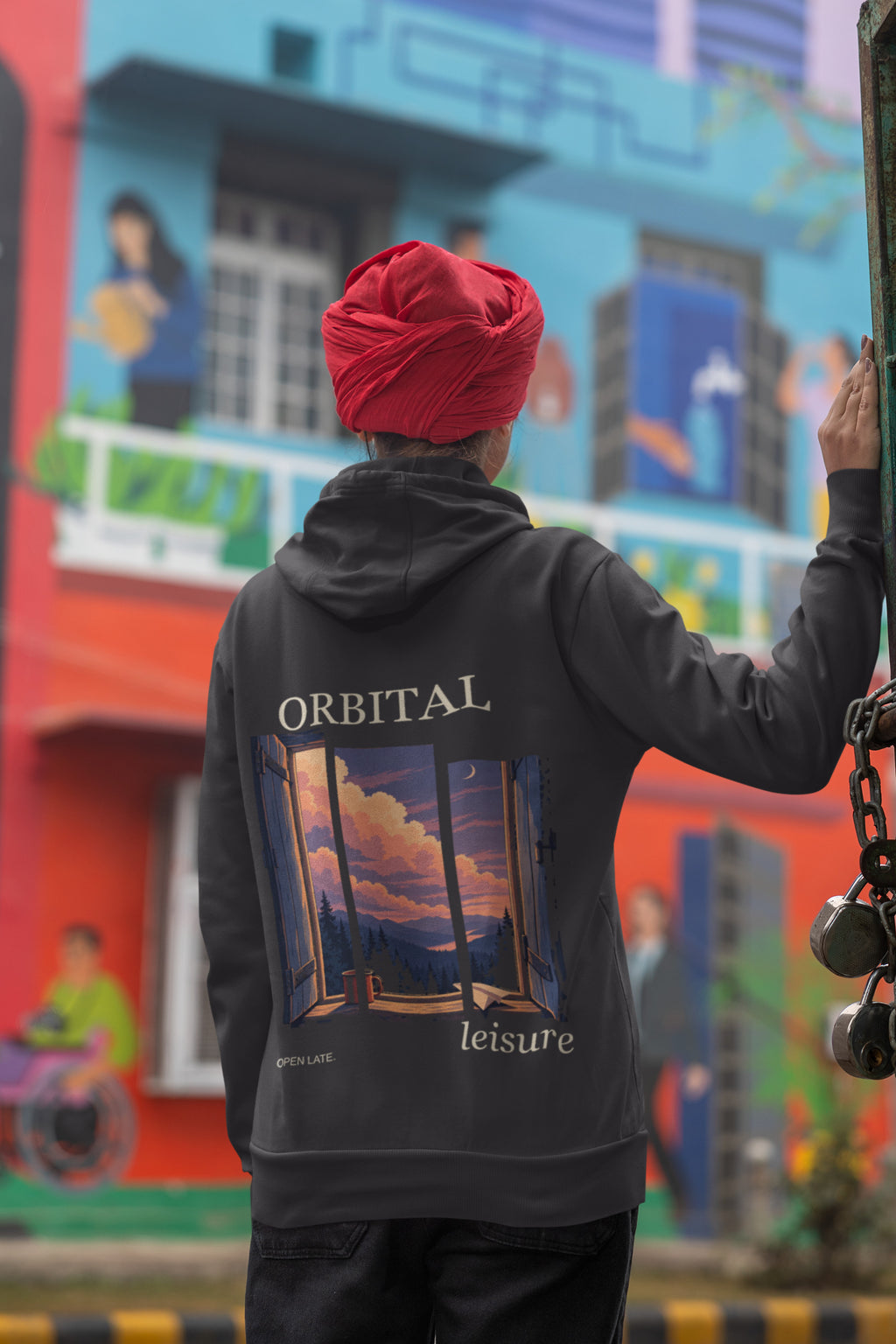 Open Late Zip Hoodie in Black, worn by a woman seen from behind in a turban by a painted mural, showing the large print on the back. Digital product mockup.
