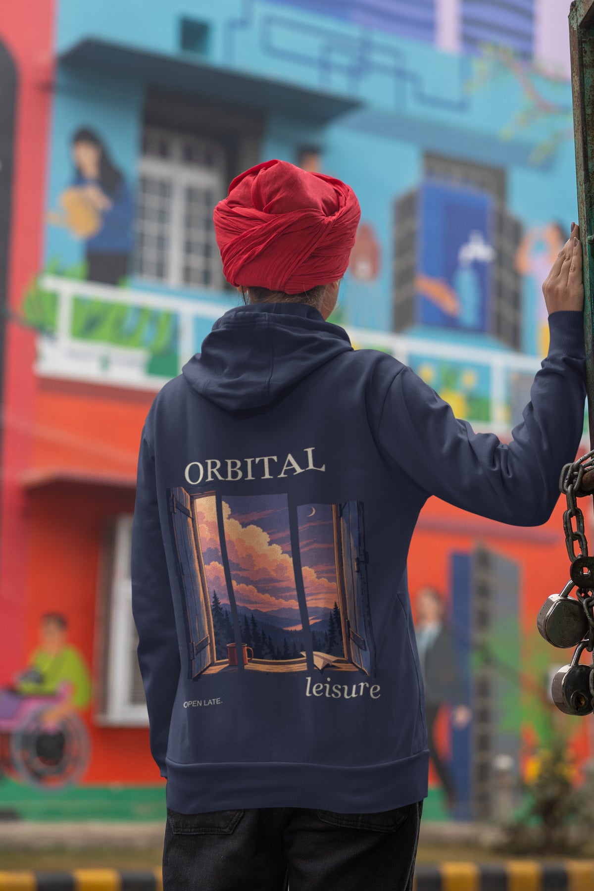 Open Late Zip Hoodie in Navy, worn by a woman seen from behind in a turban by a painted mural, showing the large print on the back. Digital product mockup.