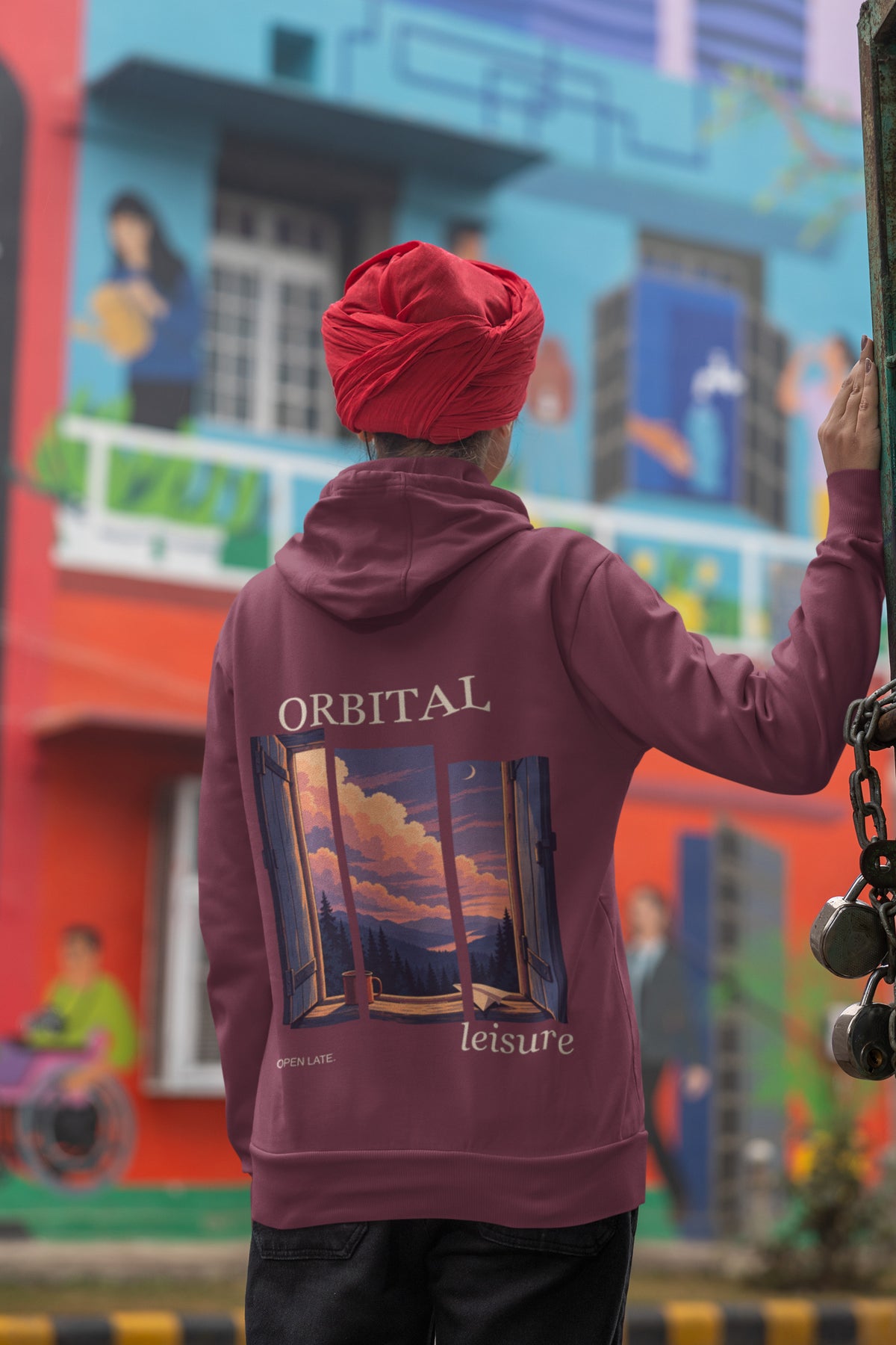 Open Late Zip Hoodie in Maroon, worn by a woman seen from behind in a turban by a painted mural, showing the large print on the back. Digital product mockup.