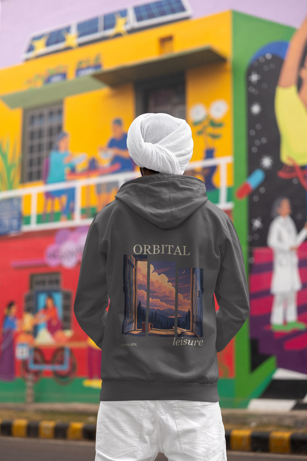 Open Late Zip Hoodie in Dark Heather, worn by a man seen from behind in a turban by a painted mural, showing the large print on the back. Digital product mockup.