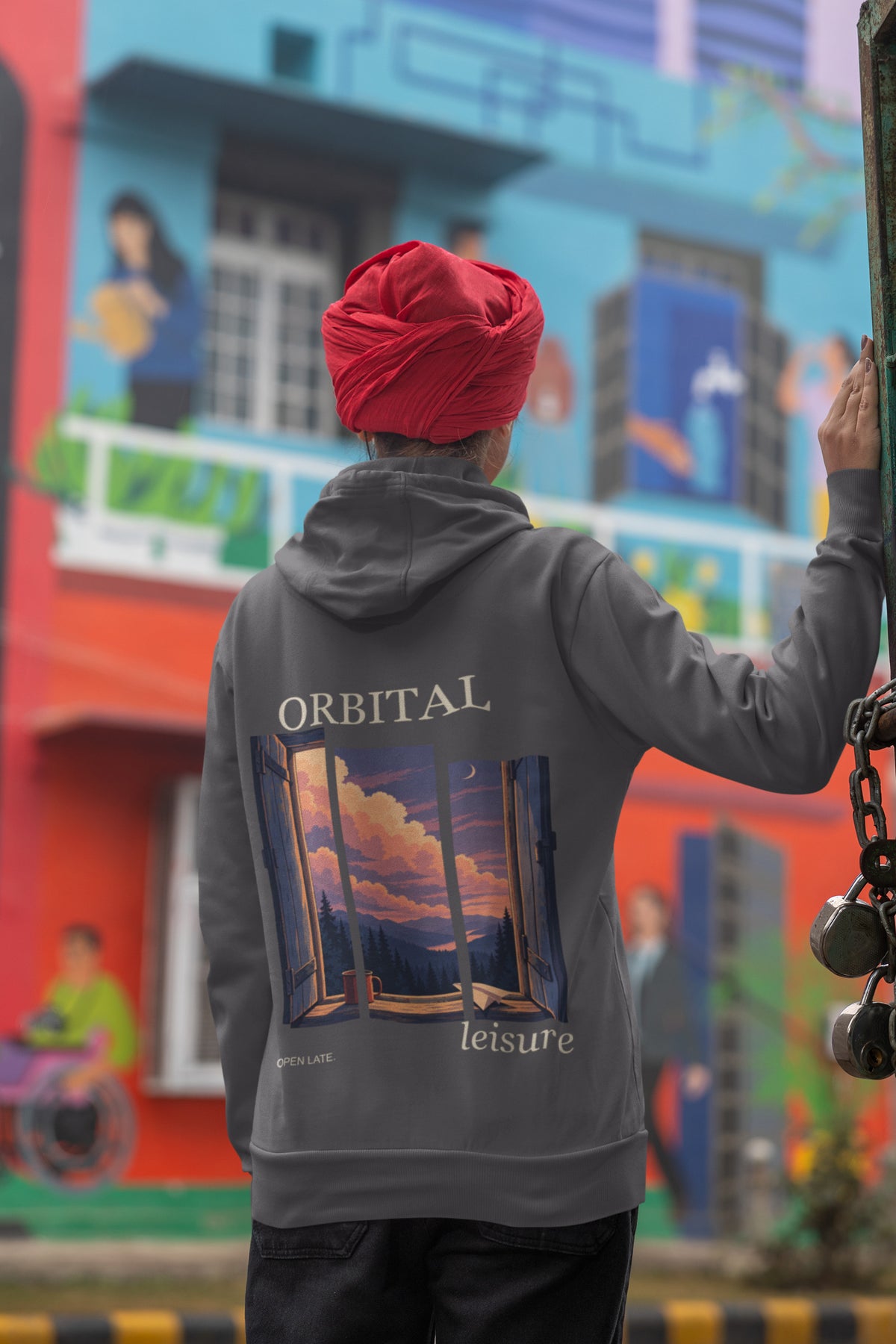 Open Late Zip Hoodie in Dark Heather, worn by a woman seen from behind in a turban by a painted mural, showing the large print on the back. Digital product mockup.
