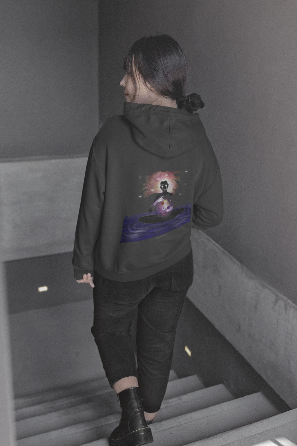 Orbit Keeper Zip Hoodie in Black, worn by a woman seen from behind walking down a staircase, showing the large print on the back. Digital product mockup.
