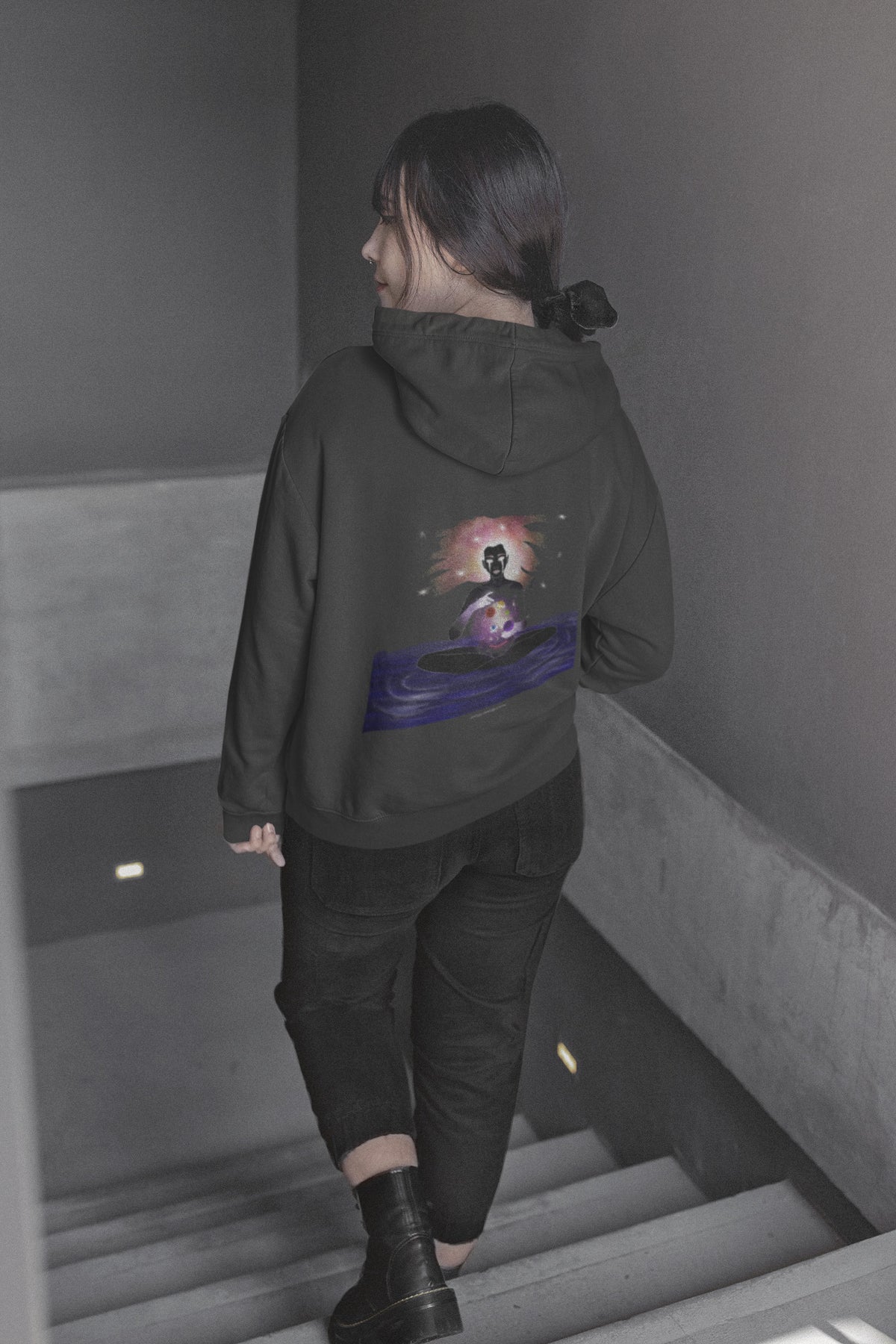 Orbit Keeper Zip Hoodie in Black, worn by a woman seen from behind walking down a staircase, showing the large print on the back. Digital product mockup.