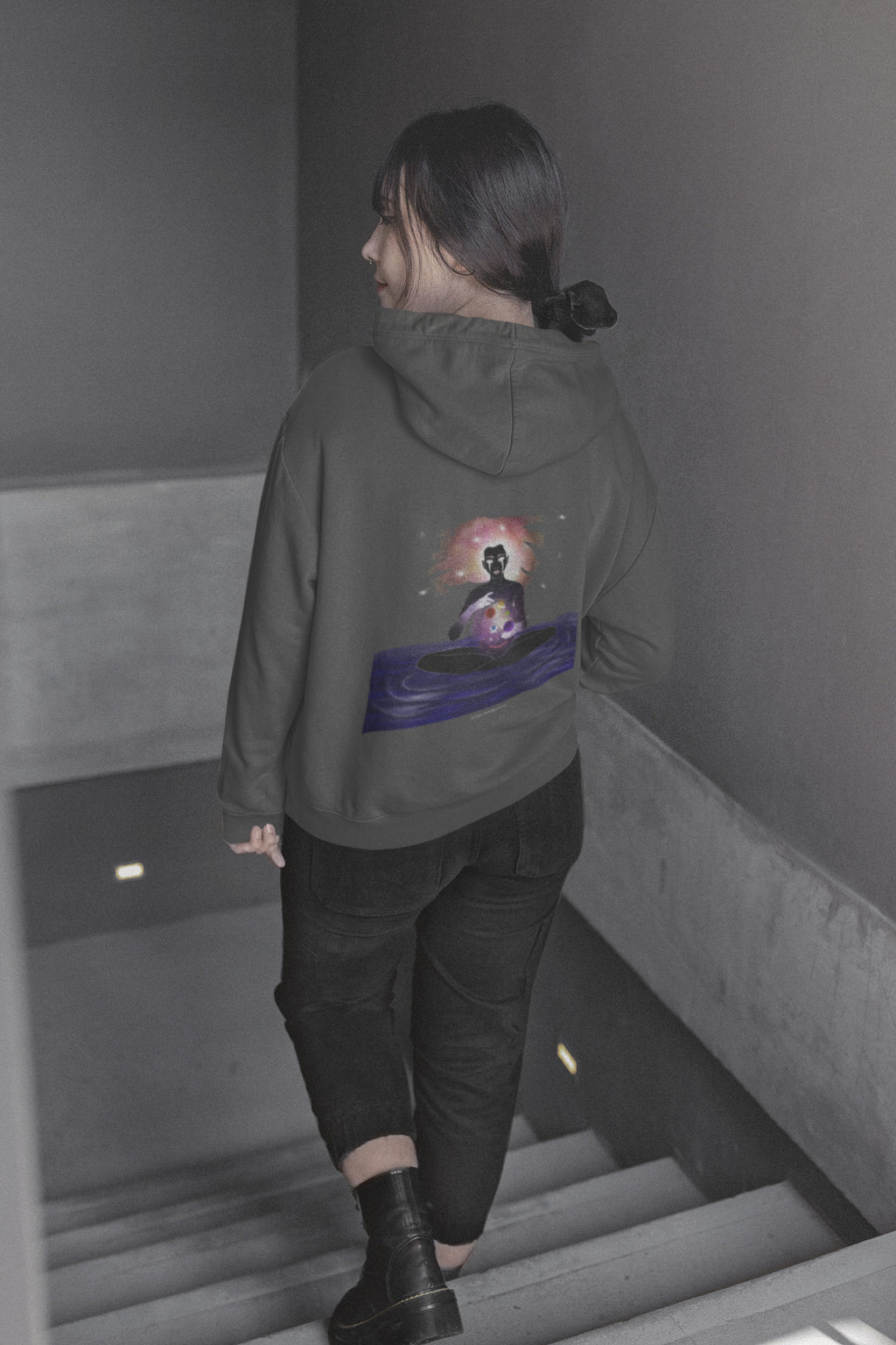 Orbit Keeper Zip Hoodie in Dark Heather, worn by a woman seen from behind walking down a staircase, showing the large print on the back. Digital product mockup.