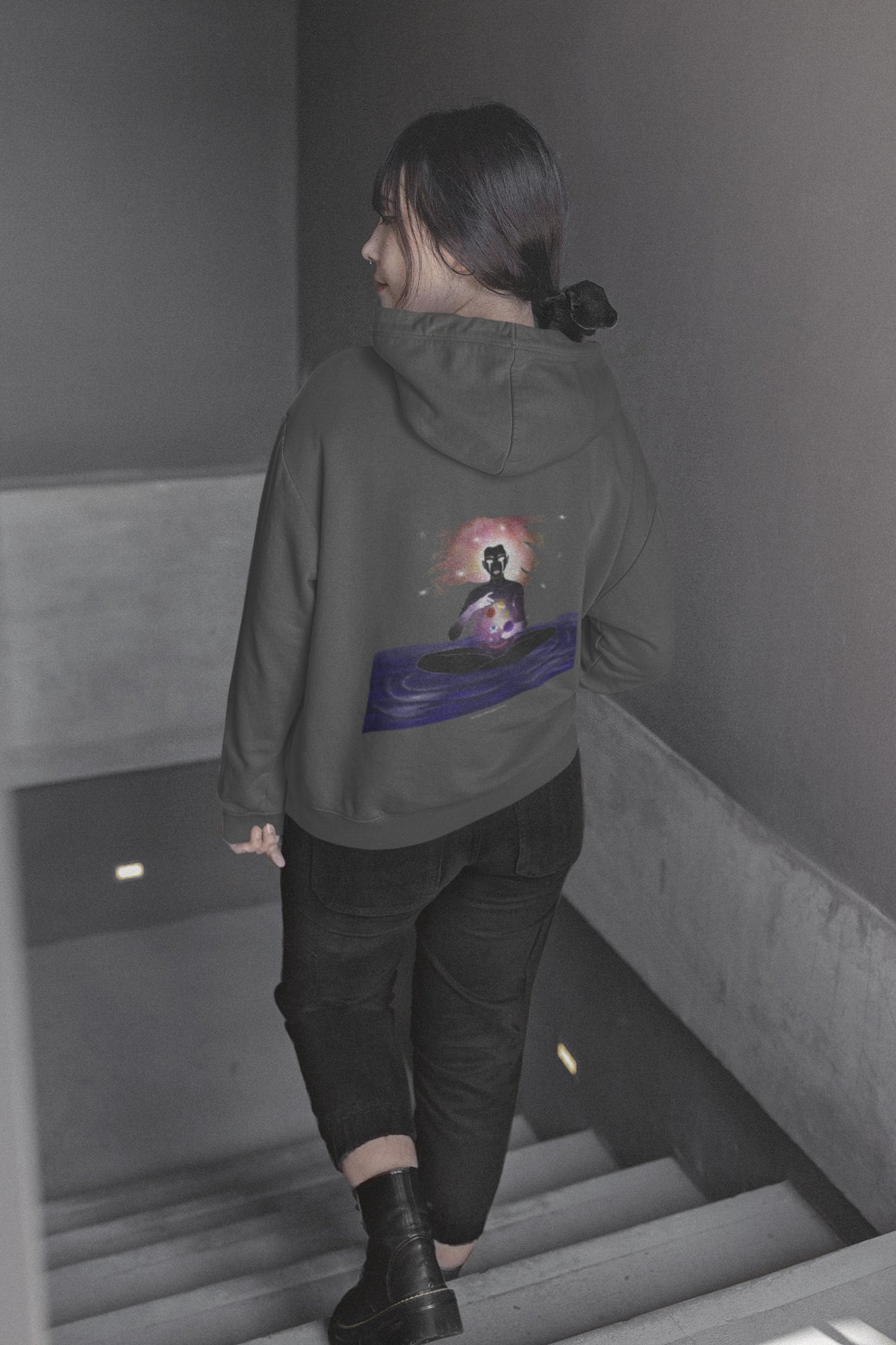 Orbit Keeper Zip Hoodie in Dark Heather, worn by a woman seen from behind walking down a staircase, showing the large print on the back. Digital product mockup.