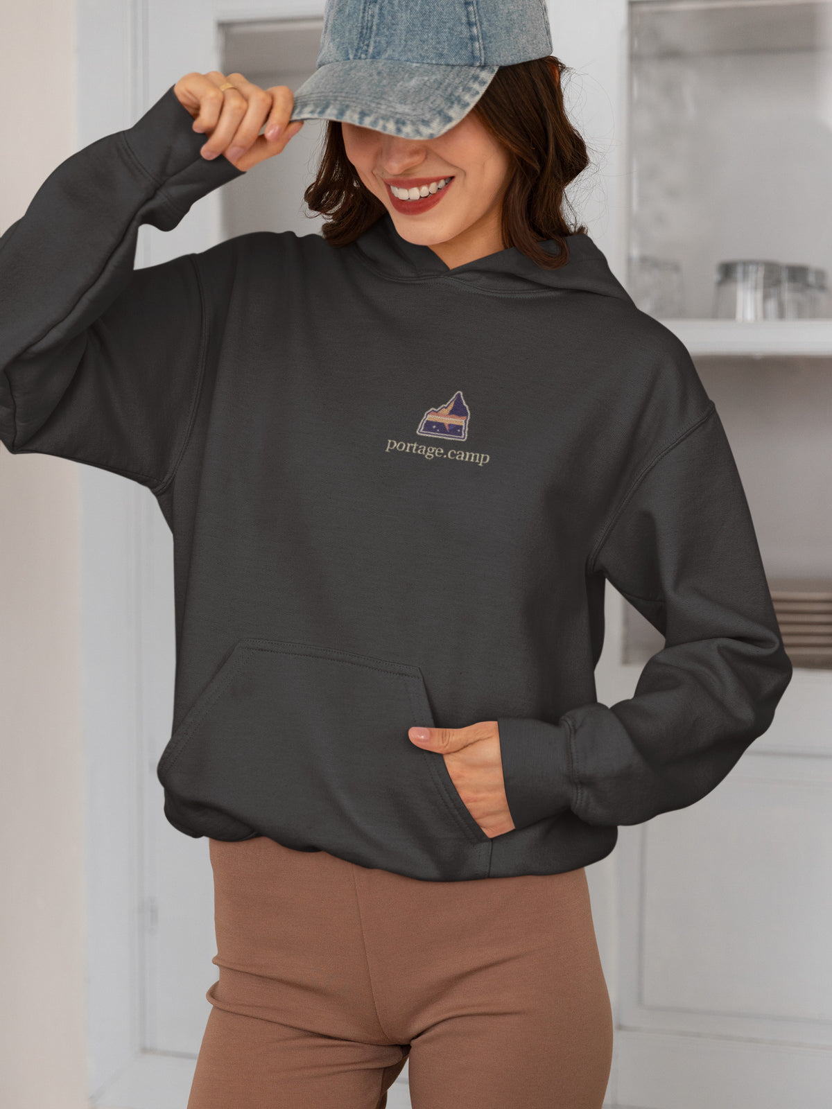 A Different Sky Heavyweight Hoodie in Black, worn by a woman smiling in a kitchen, with the A Different Sky logo and portage.camp on the left chest. Digital product mockup.