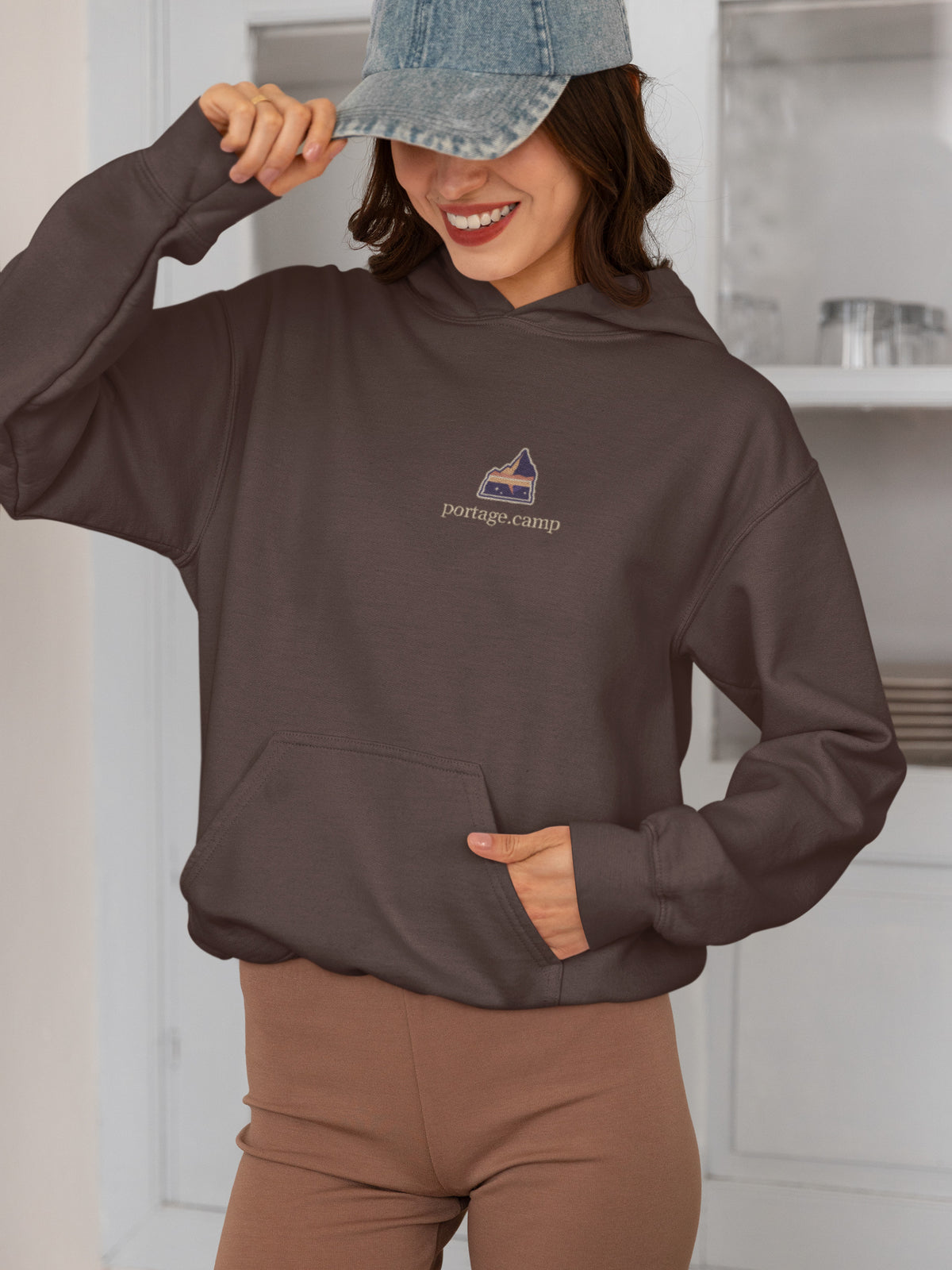 A Different Sky Heavyweight Hoodie in Brown, worn by a woman smiling in a kitchen, with the A Different Sky logo and portage.camp on the left chest. Digital product mockup.
