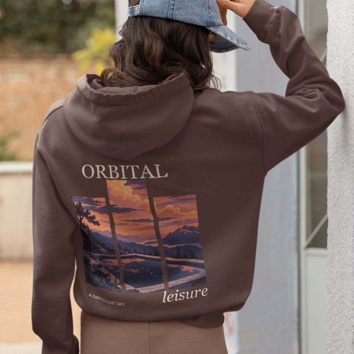 A Different Sky Heavyweight Hoodie in Brown, worn by a woman seen from behind at home, showing the large print on the back. Digital product mockup.
