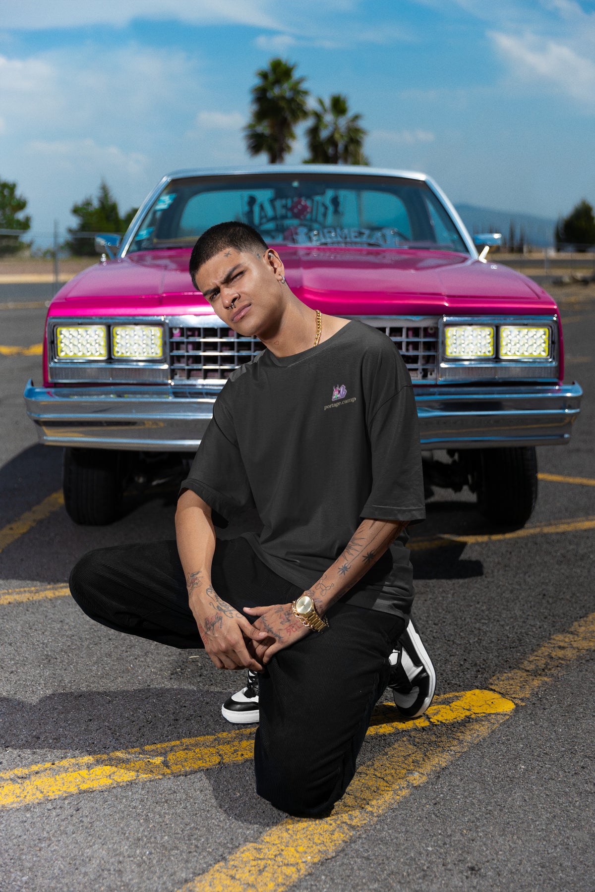 Amethyst After Hours Garment-Dyed Tee in Black, worn by a man crouched in front of a pink classic car, with the Amethyst After Hours logo and portage.camp on the chest. Digital product mockup.