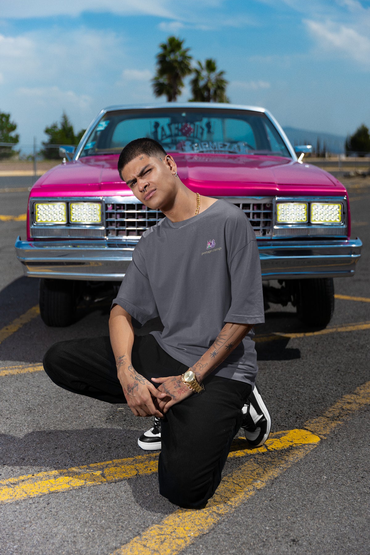 Amethyst After Hours Garment-Dyed Tee in Navy, worn by a man crouched in front of a pink classic car, with the Amethyst After Hours logo and portage.camp on the chest. Digital product mockup.