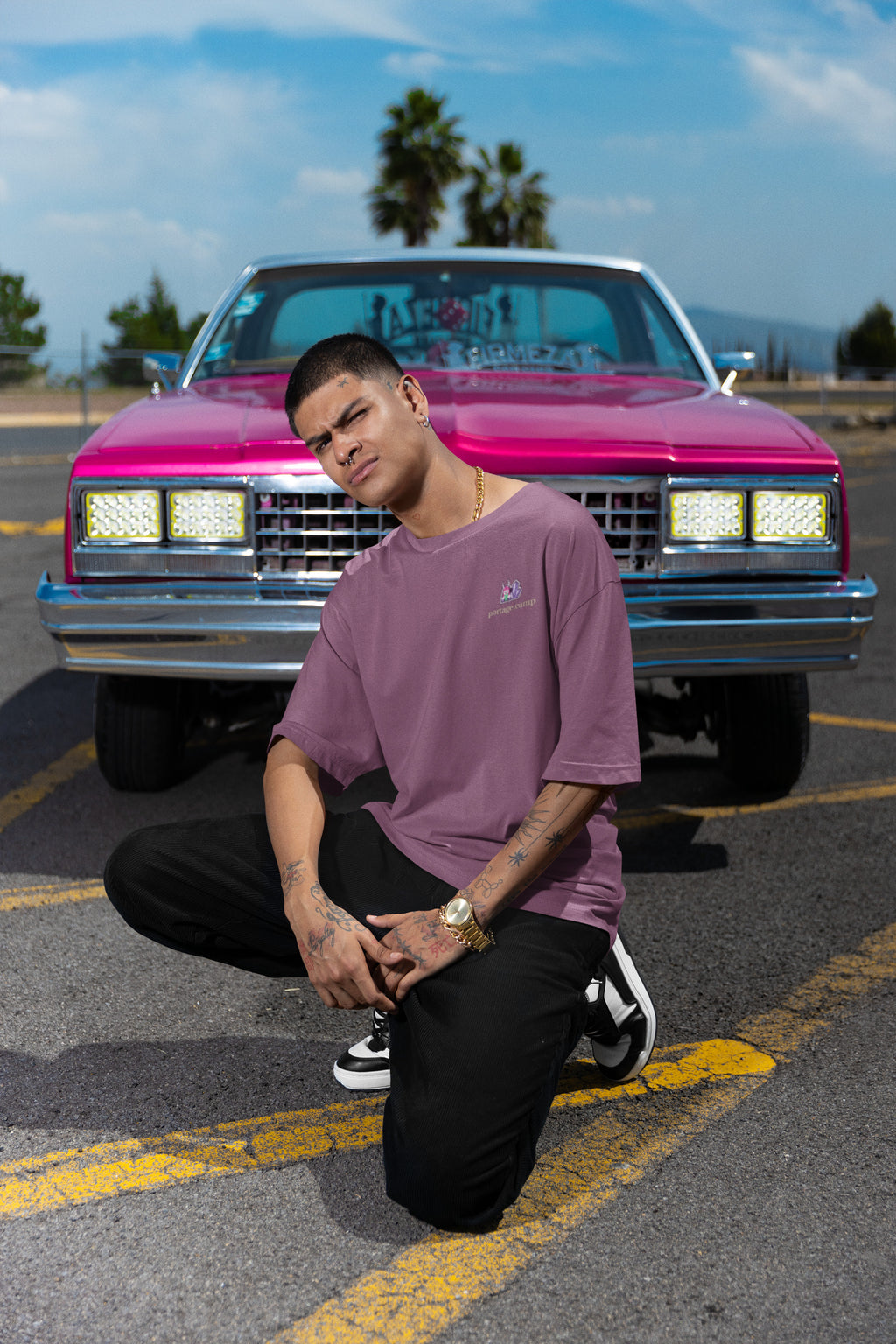 Amethyst After Hours Garment-Dyed Tee in Berry, worn by a man crouched in front of a pink classic car, with the Amethyst After Hours logo and portage.camp on the chest. Digital product mockup.