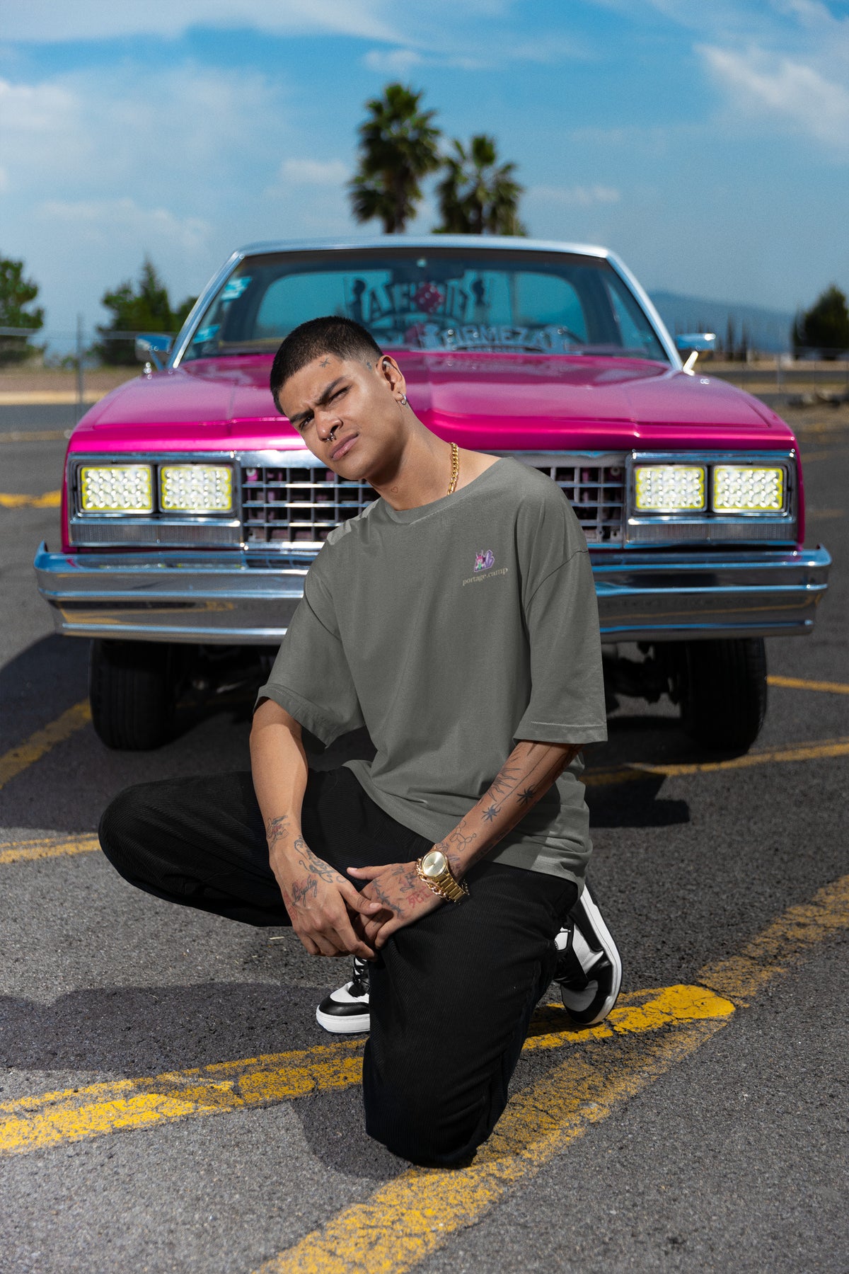 Amethyst After Hours Garment-Dyed Tee in Pepper, worn by a man crouched in front of a pink classic car, with the Amethyst After Hours logo and portage.camp on the chest. Digital product mockup.