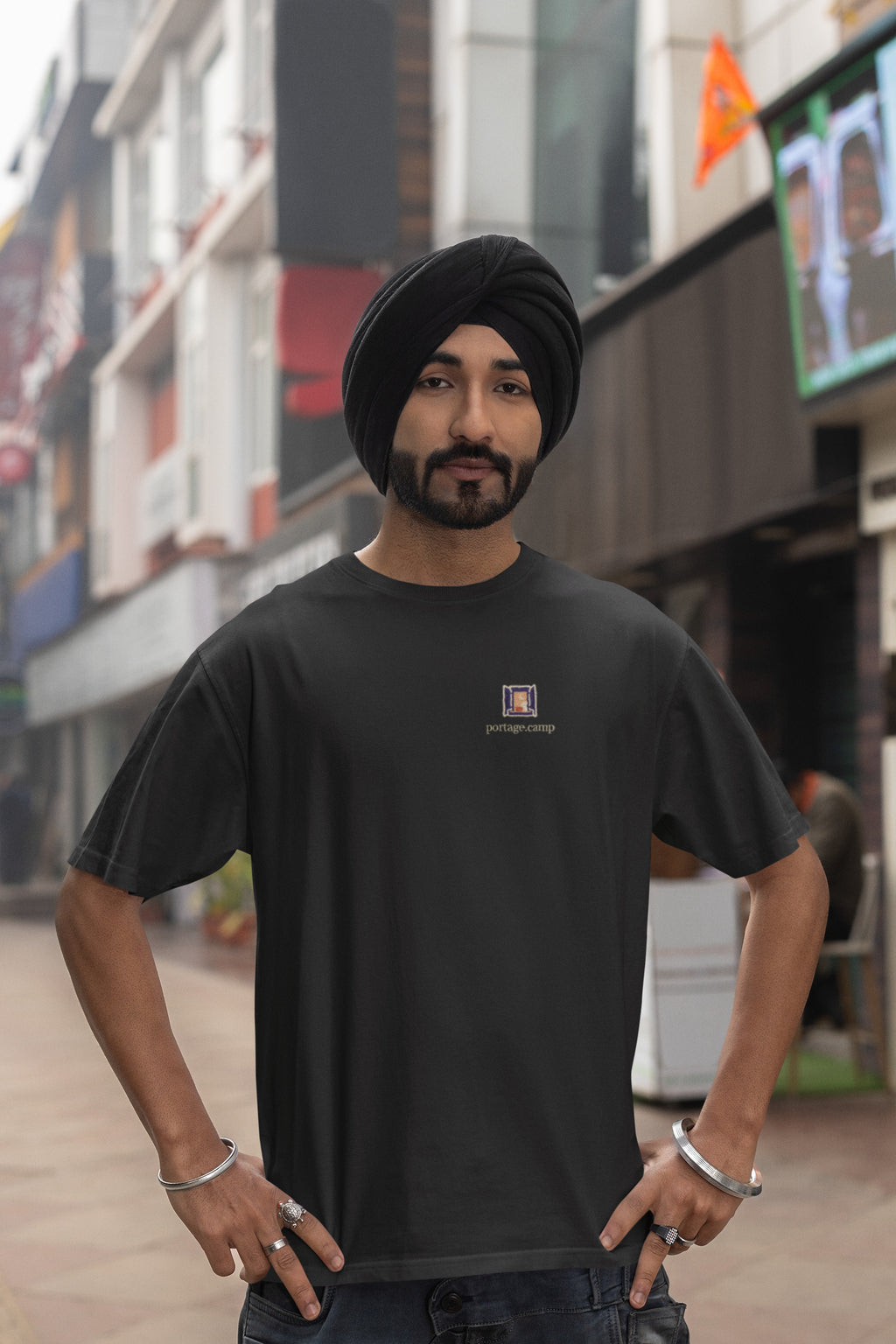 Open Late Garment-Dyed Tee in Black, worn by a man in a turban on a busy street, with the Open Late logo and portage.camp on the chest. Digital product mockup.