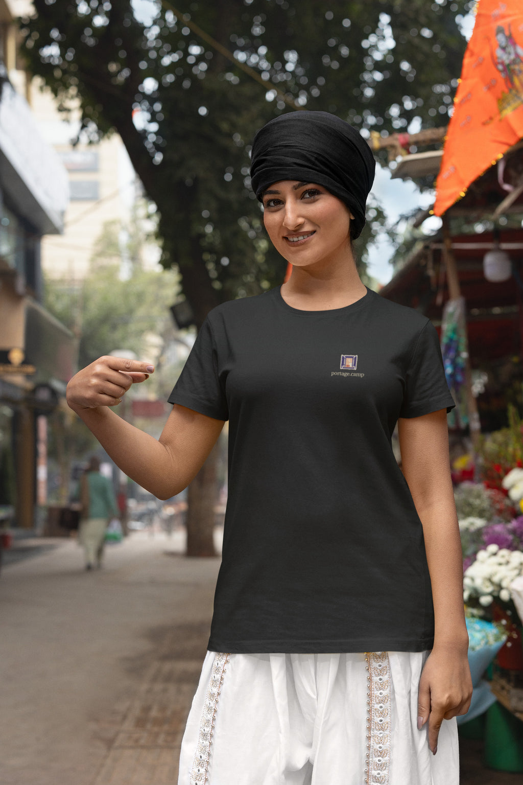 Open Late Garment-Dyed Tee in Black, worn by a woman in a turban by a flower market, with the Open Late logo and portage.camp on the chest. Digital product mockup.