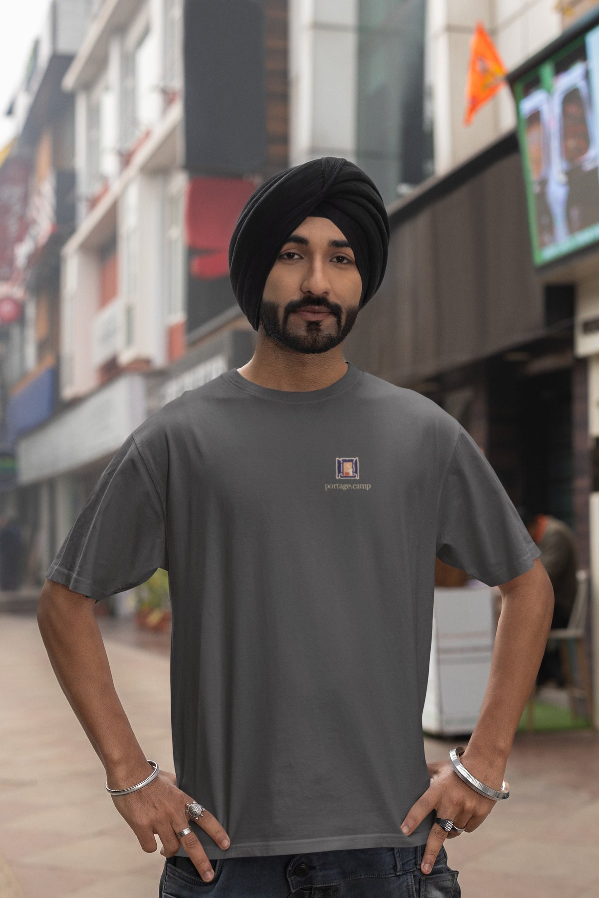 Open Late Garment-Dyed Tee in Graphite, worn by a man in a turban on a busy street, with the Open Late logo and portage.camp on the chest. Digital product mockup.